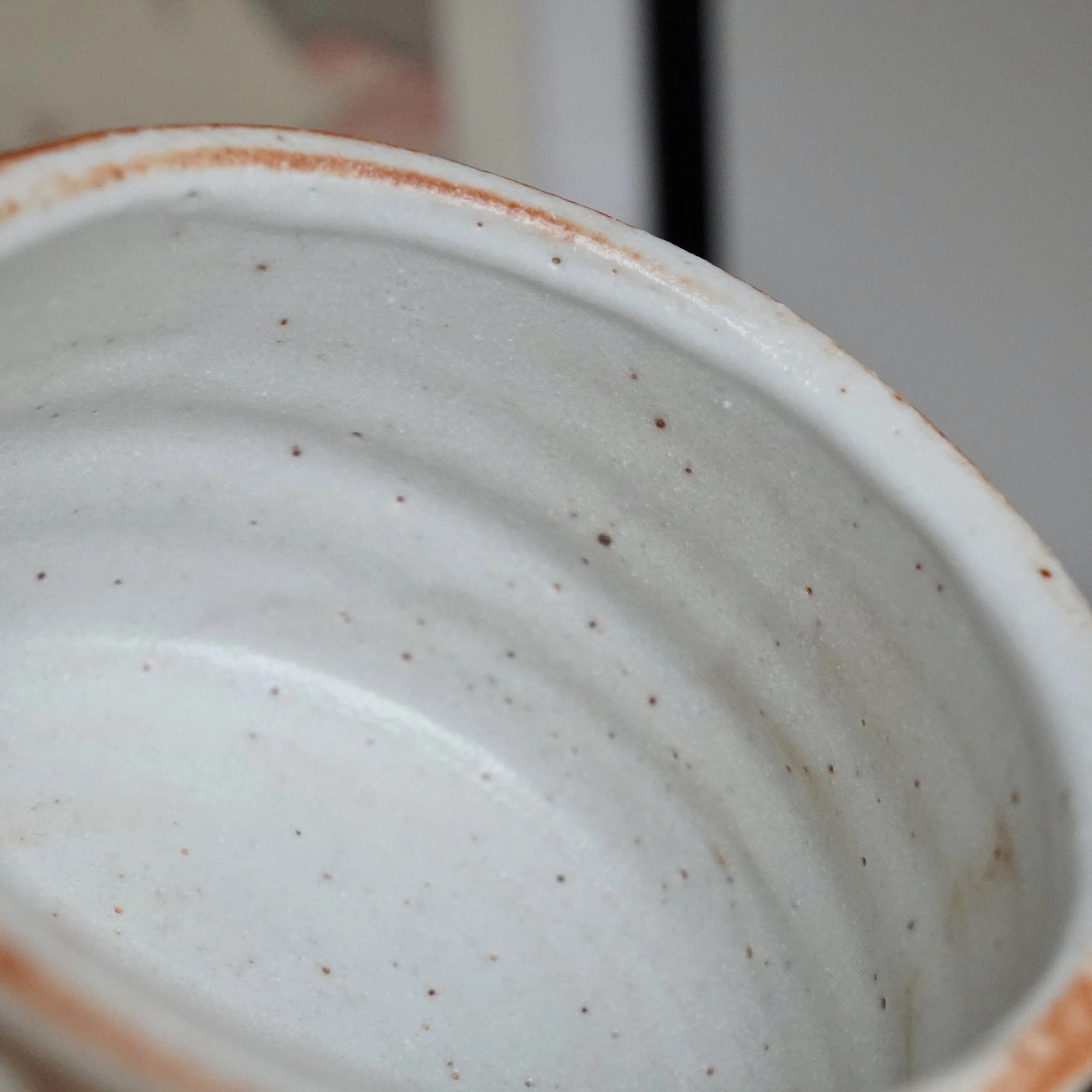 Interior of a Shino chawan, white glazed with a rust-orange blush on its irregular rim and small brown speckles throughout.