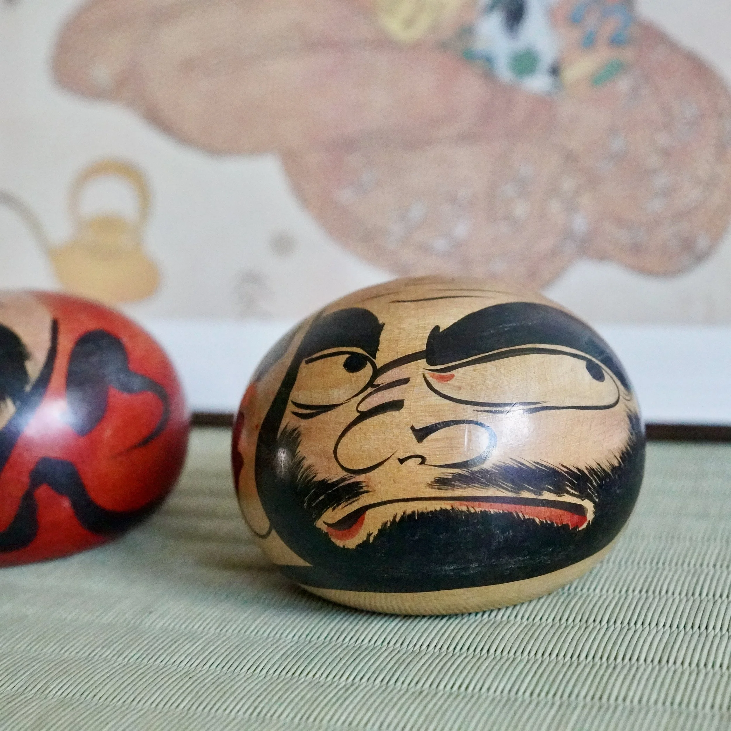 Close-up of a hand-painted wooden Tsugaru Daruma doll by Satō Zenji, featuring a scowling face with black and red details, next to a blurred red doll on a woven mat.