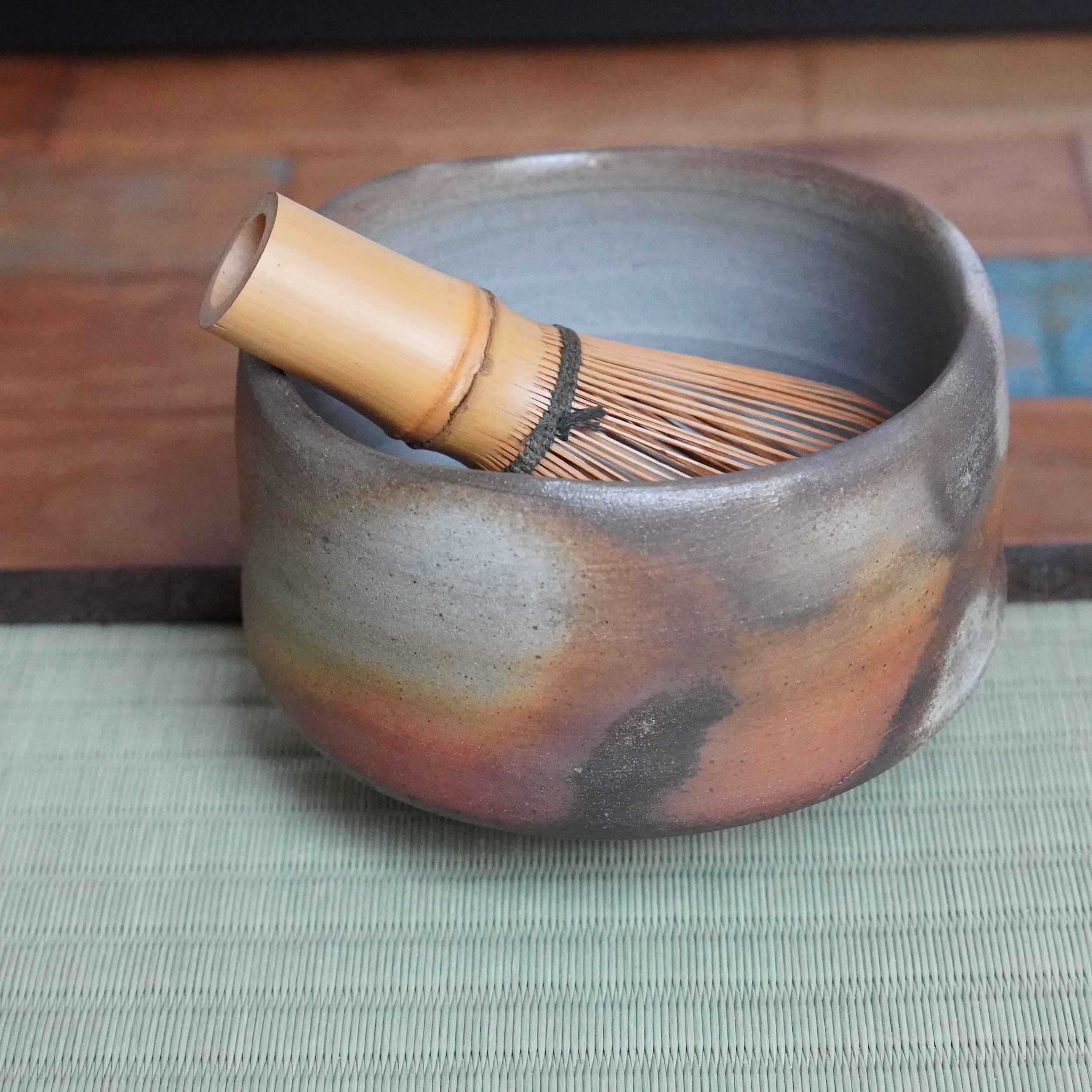 Inomata Masaaki Bizen chawan, fired with tones of deep rust, burnt orange, and ash grey, holds a bamboo tea whisk on a woven mat.