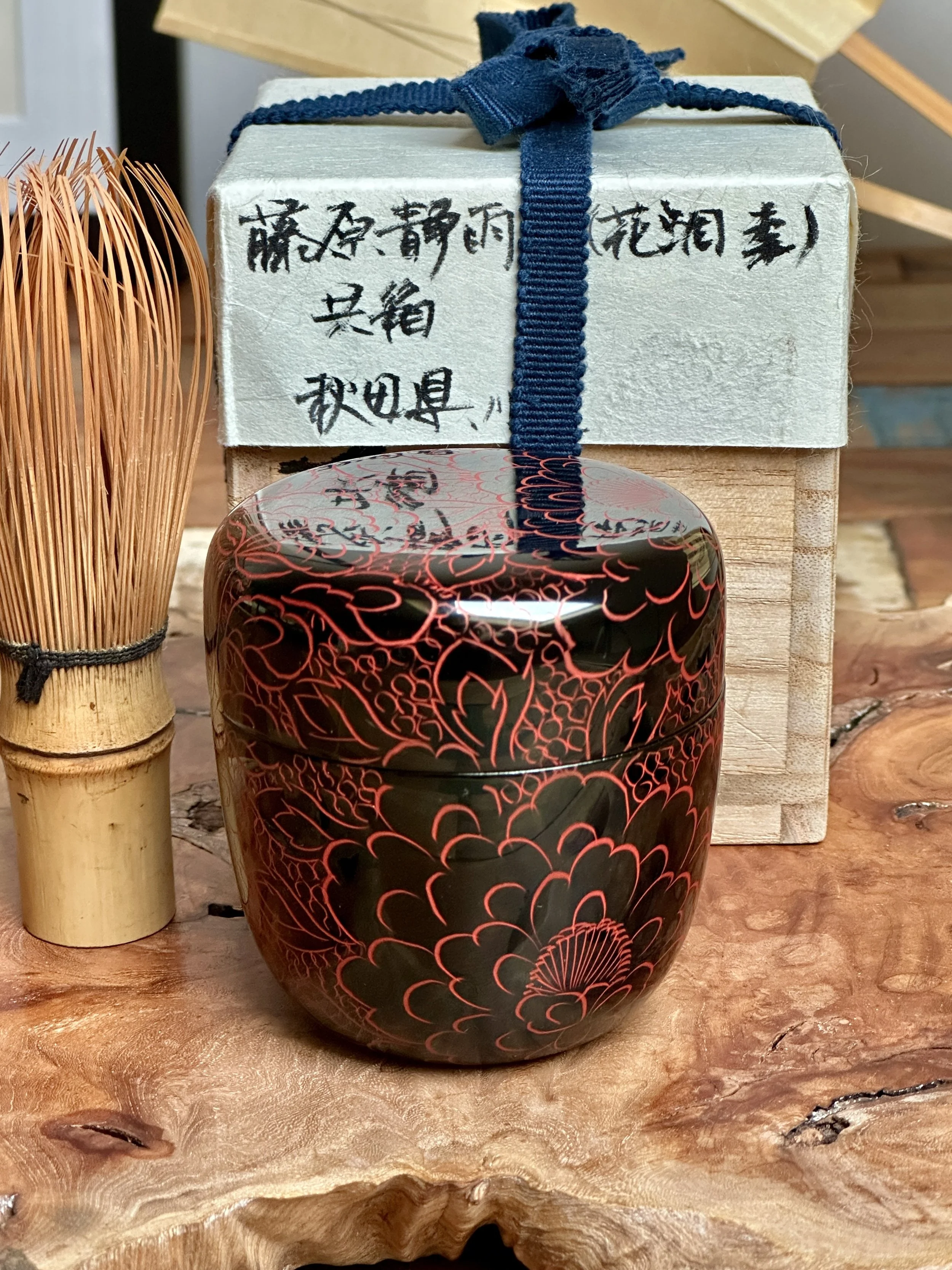 Black lacquered tea caddy with engraved vermilion peony design, bamboo whisk, and wooden box signed Fujiwara Seiu, on a textured wood surface.