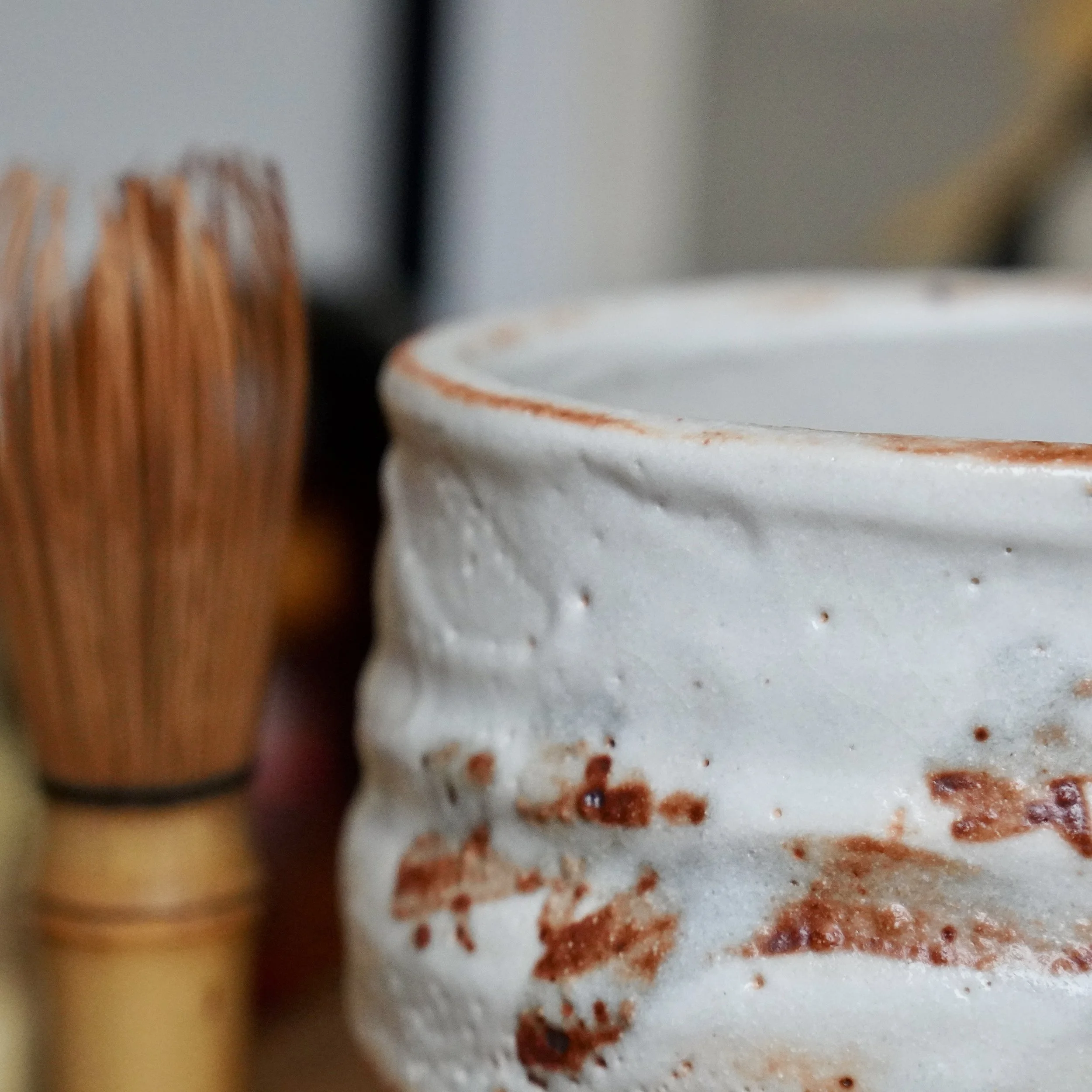 Shino chawan with textured white glaze and irregular rust-orange markings; a blurred bamboo tea whisk in the background.