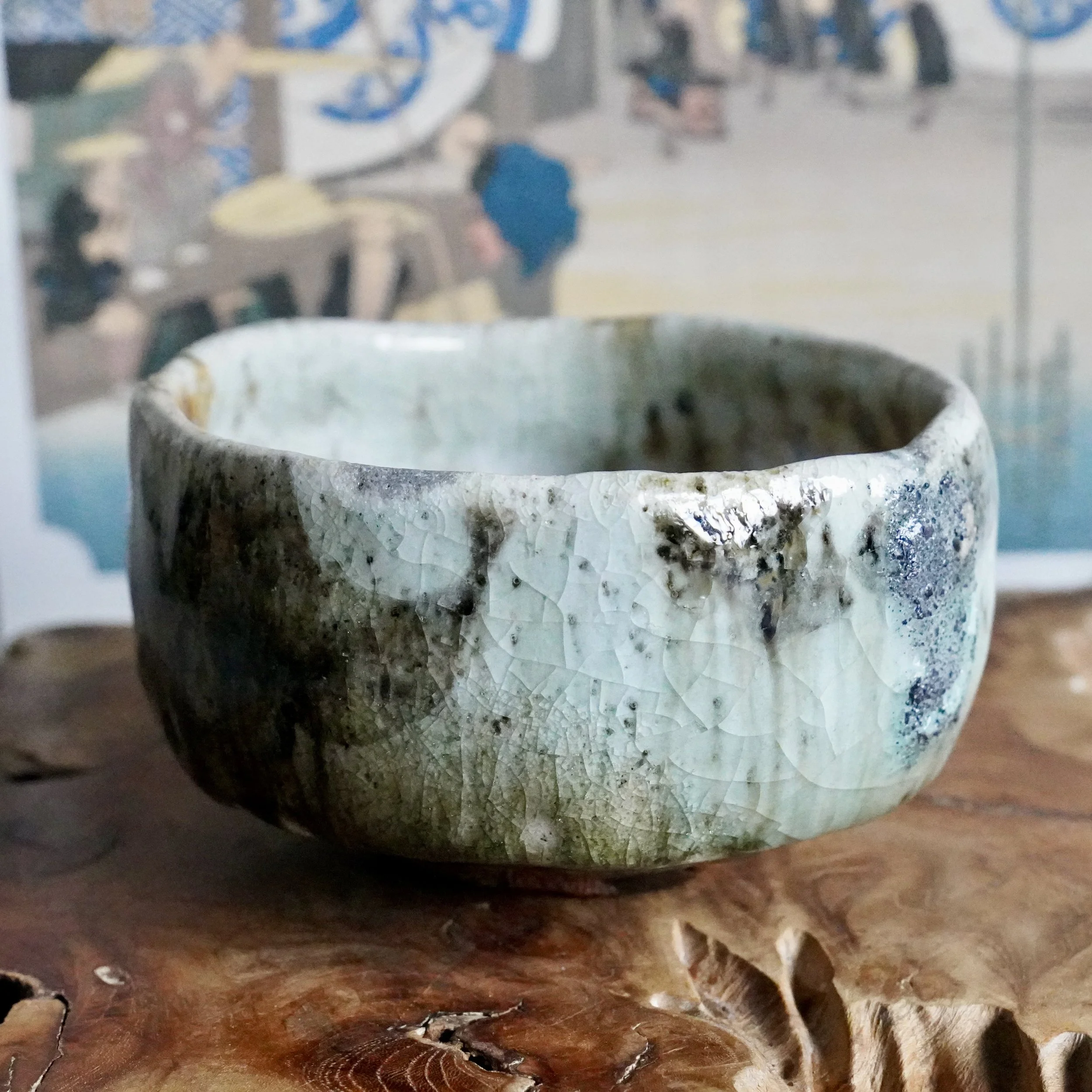 Deguchi Eiji chawan with heavily crackled glaze in pale green, dark brown, and a blue-grey patch, on a wood surface.