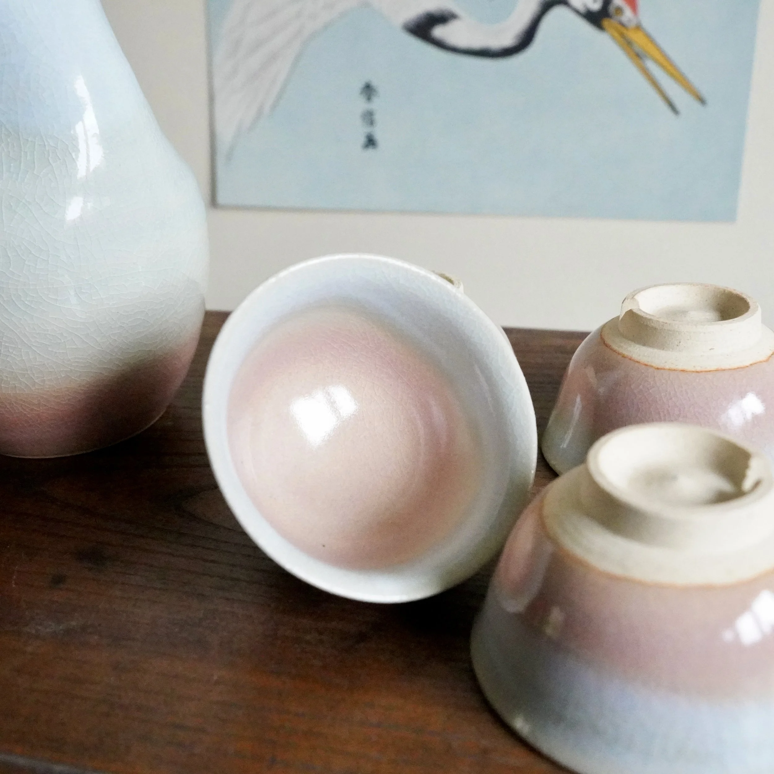 Hagi-yaki sake set: a tokkuri and three ochoko with gradient pink to pale blue-grey crazed glaze, on wood with a background bird print.