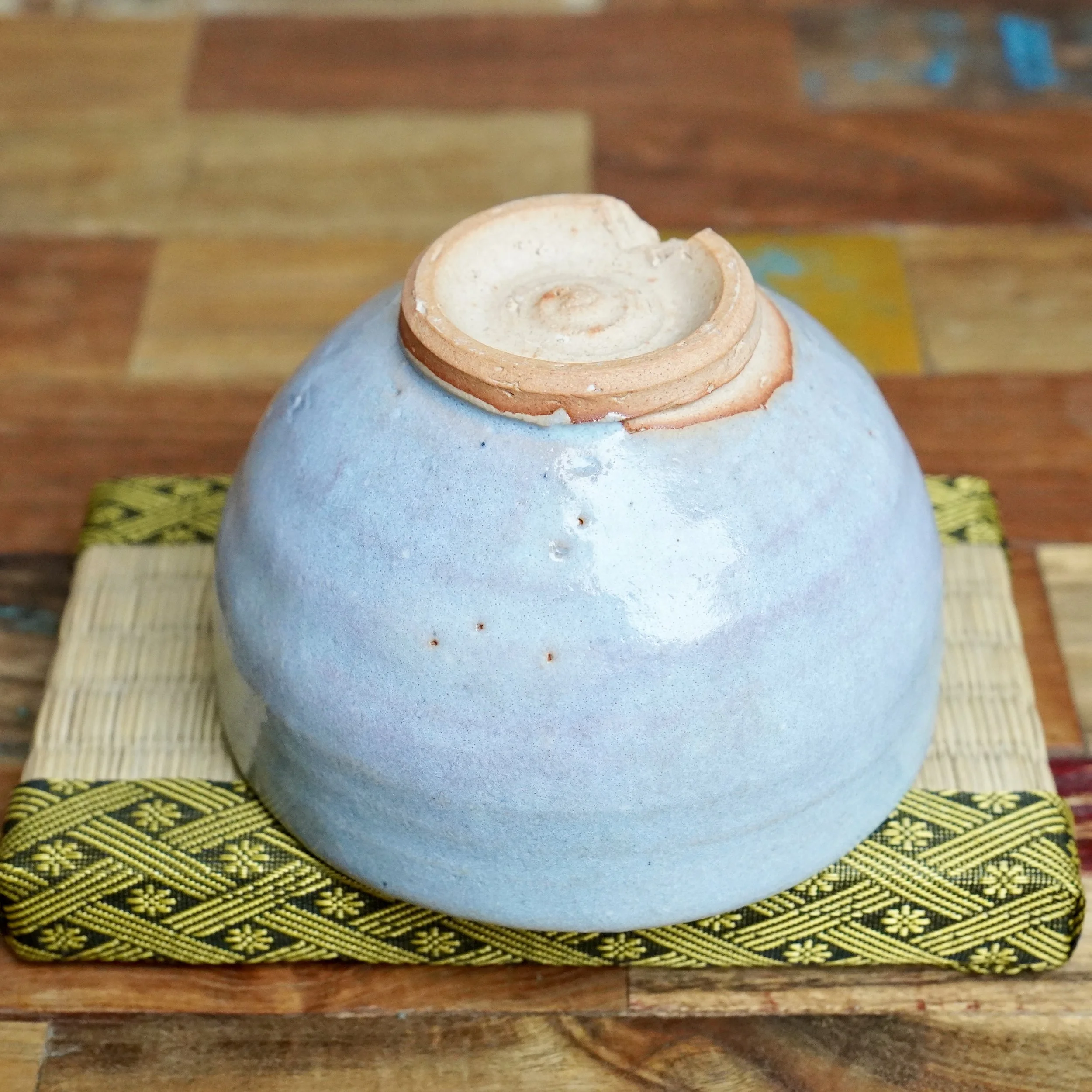 Moriwaki Fumitada Hagi-yaki chawan, light blue-grey with textured glaze and small spots, rests upside down, revealing its unglazed reddish-brown warikodai with spiral mark and chipped edge.