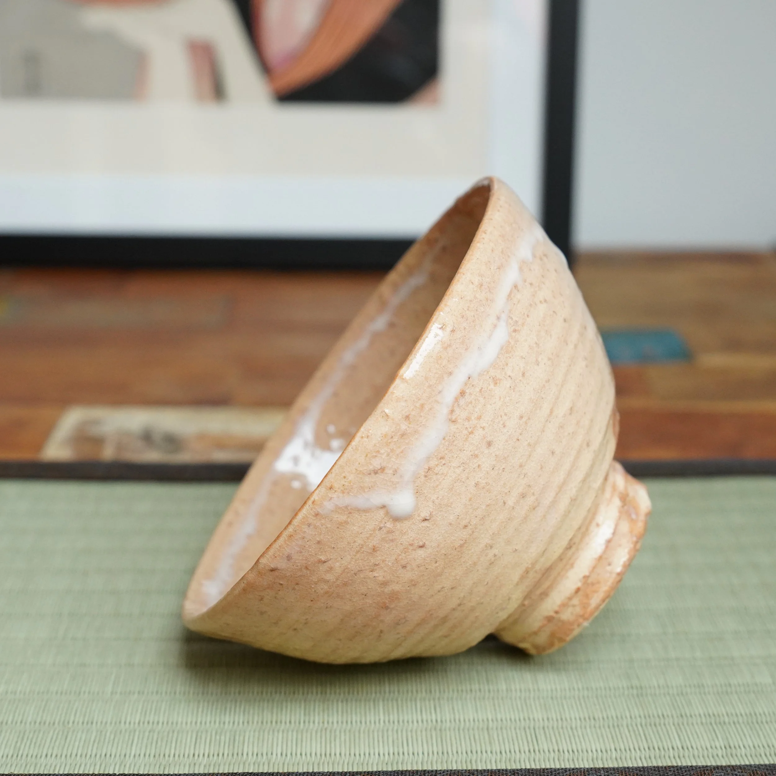 Kaneko Nobuhiko's Hagi-yaki chawan, tilted on a green tatami mat, shows a salmon-pink body with wheel rings and a prominent white glaze drip. Blurred art and wood are in the background.