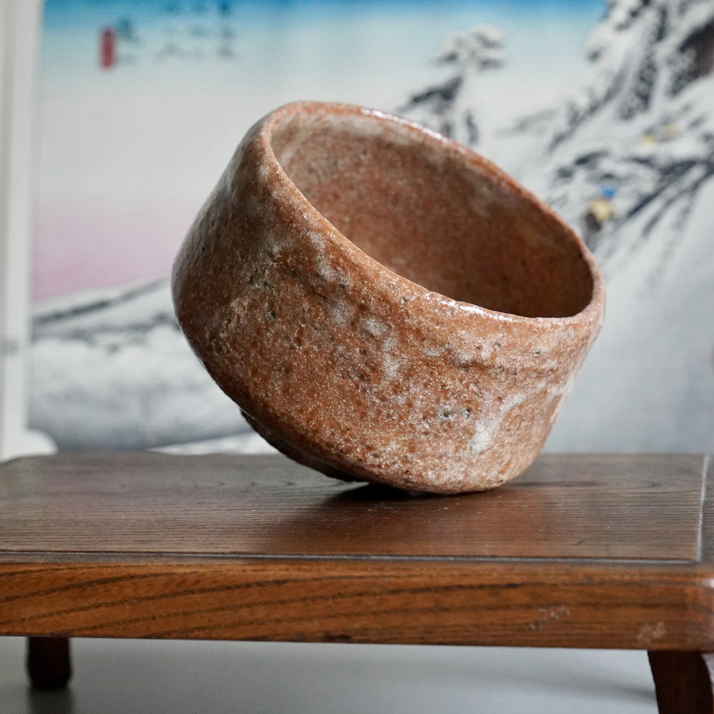 Sawada Hiroyuki aka-raku chawan, reddish-brown and textured with white glaze runs, on a dark wooden stand.