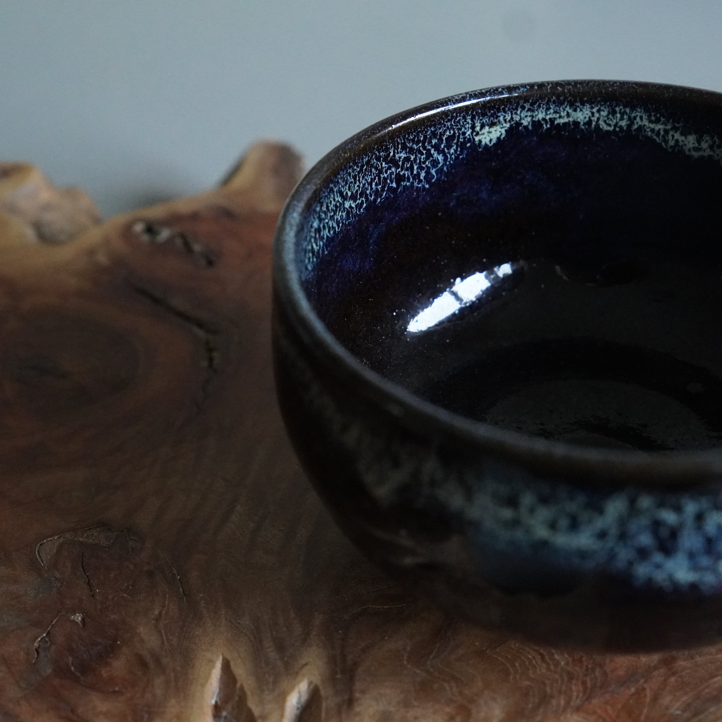 Close-up of a dark brown matcha chawan by Rokuhō, with a speckled blue and white glaze pattern on its rim and interior, on a textured wooden surface.