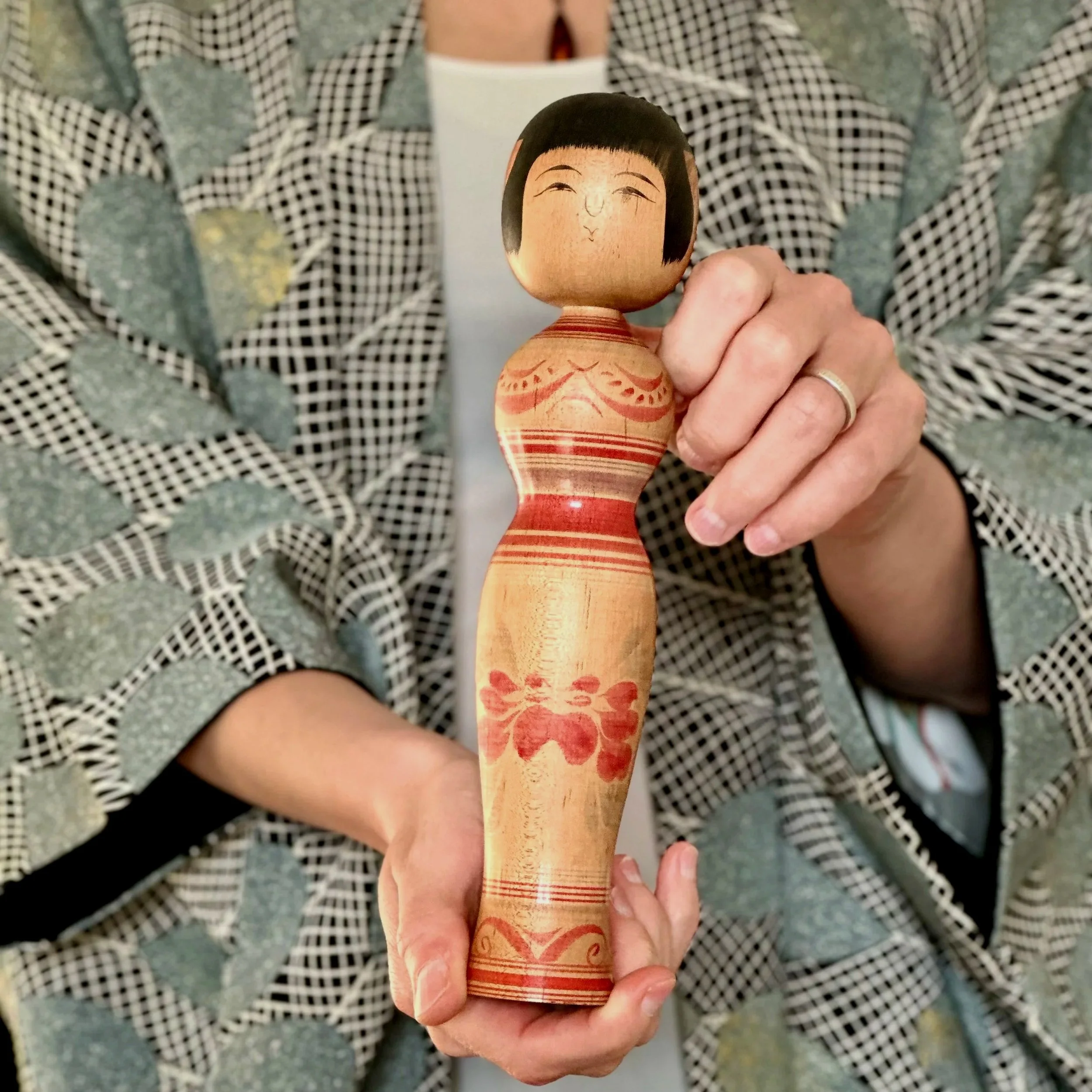 Wooden Tsugaru kokeshi doll, hourglass shaped, with red peony motif and painted face, held by hands against a patterned kimono. Sasamori Junichi.