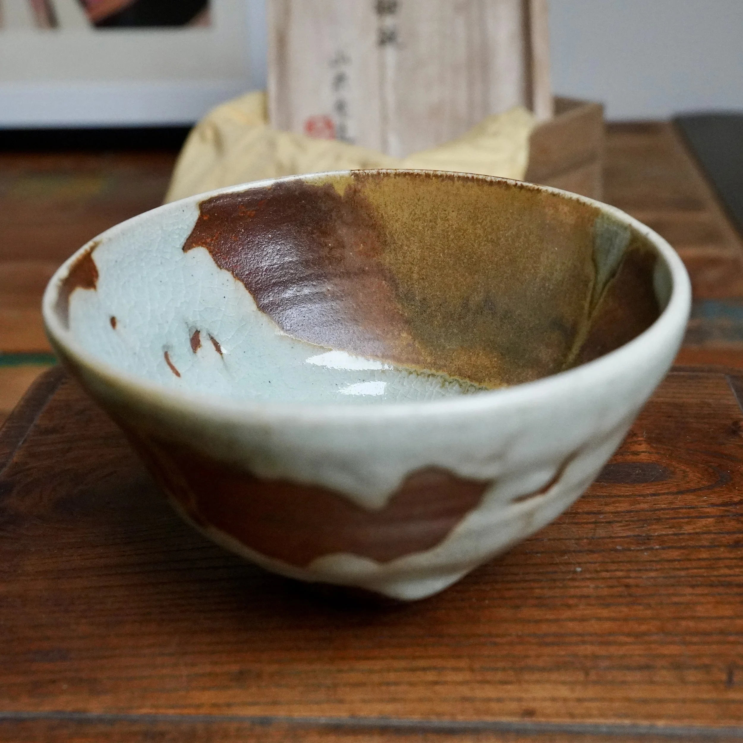 Chawan with pale crackled blue-grey glaze and flowing rust-brown and ochre, on wood with a blurred signed Sansōan tomobako in the background.