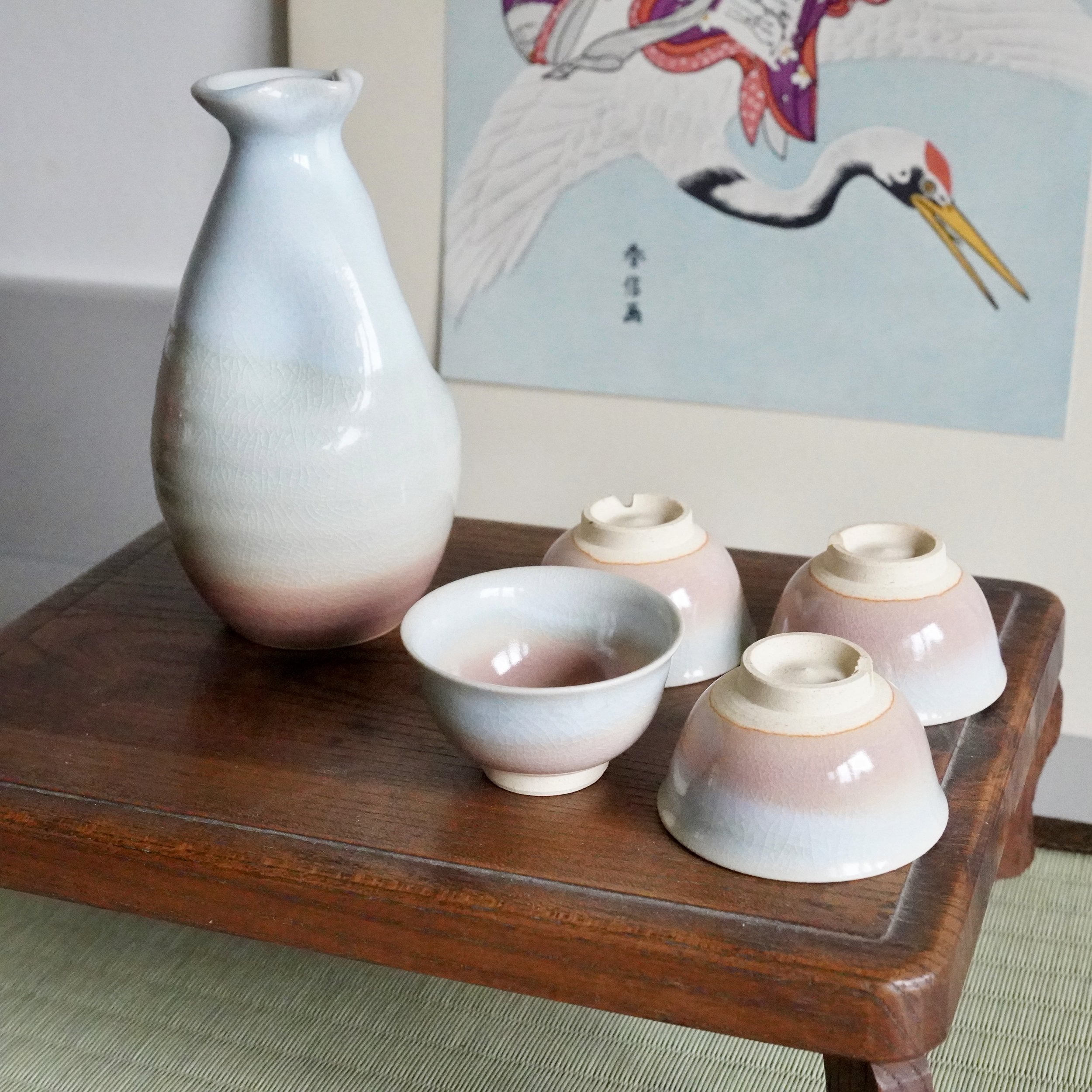 Hagi-yaki sake set: a tokkuri and four ochoko with blush-to-blue gradient glaze and crazing. They sit on a dark wooden table; one ochoko is upright, others inverted.