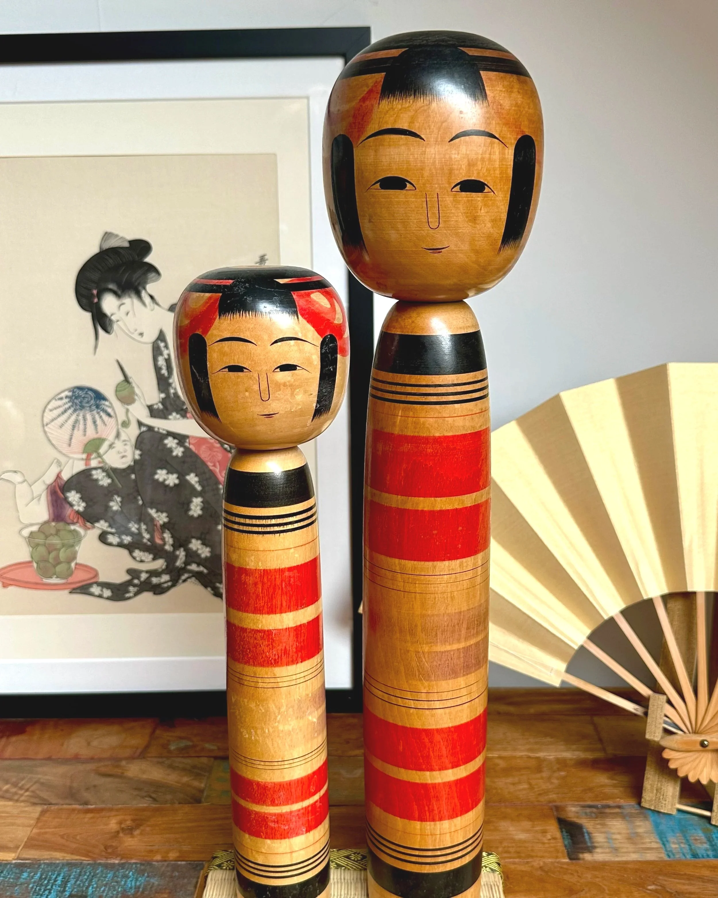 Two Tsuchiyu kokeshi dolls by Abe Ichirō, one tall, one shorter, stand on wood, featuring red and black bands, painted faces.