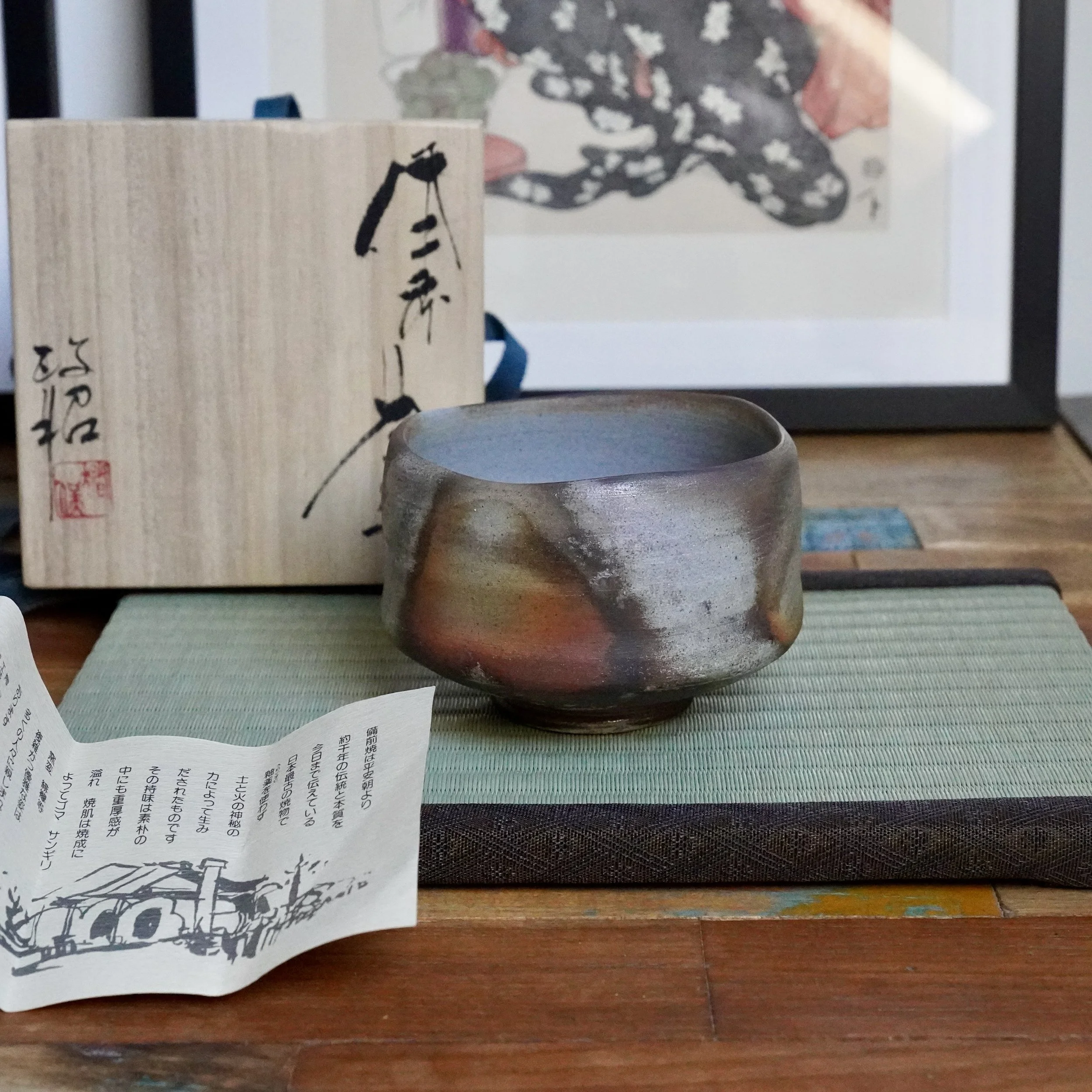 Irregularly shaped Sangiri Bizen chawan by Inomata Masaaki, showing unglazed rust, orange, ash grey, and silver tones, on a tatami mat next to its signed wooden box.