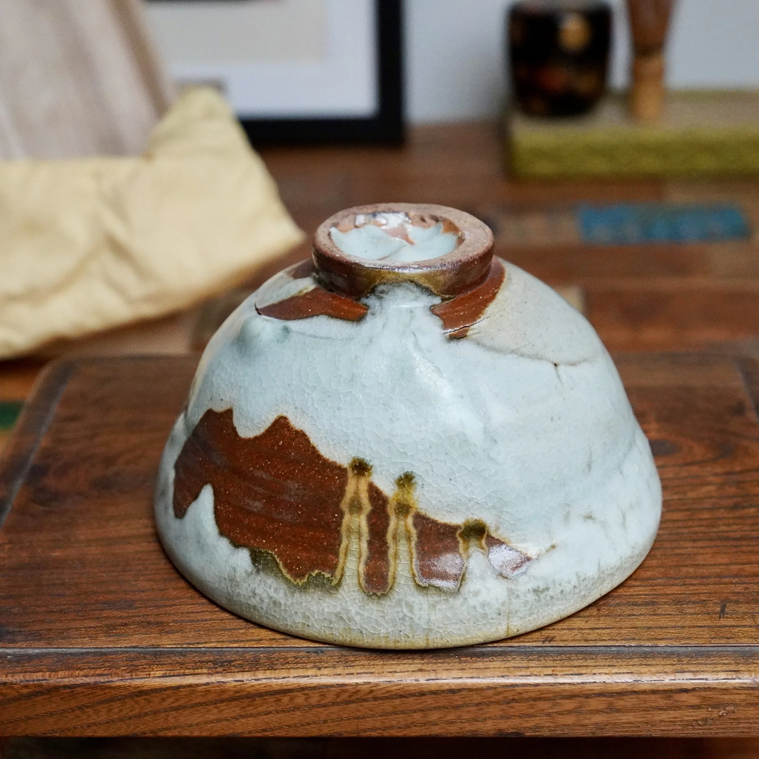 Sansōan chawan inverted on wood, featuring a pale blue-grey crackle glaze with flowing rust-brown and ochre streaks, unglazed foot visible.