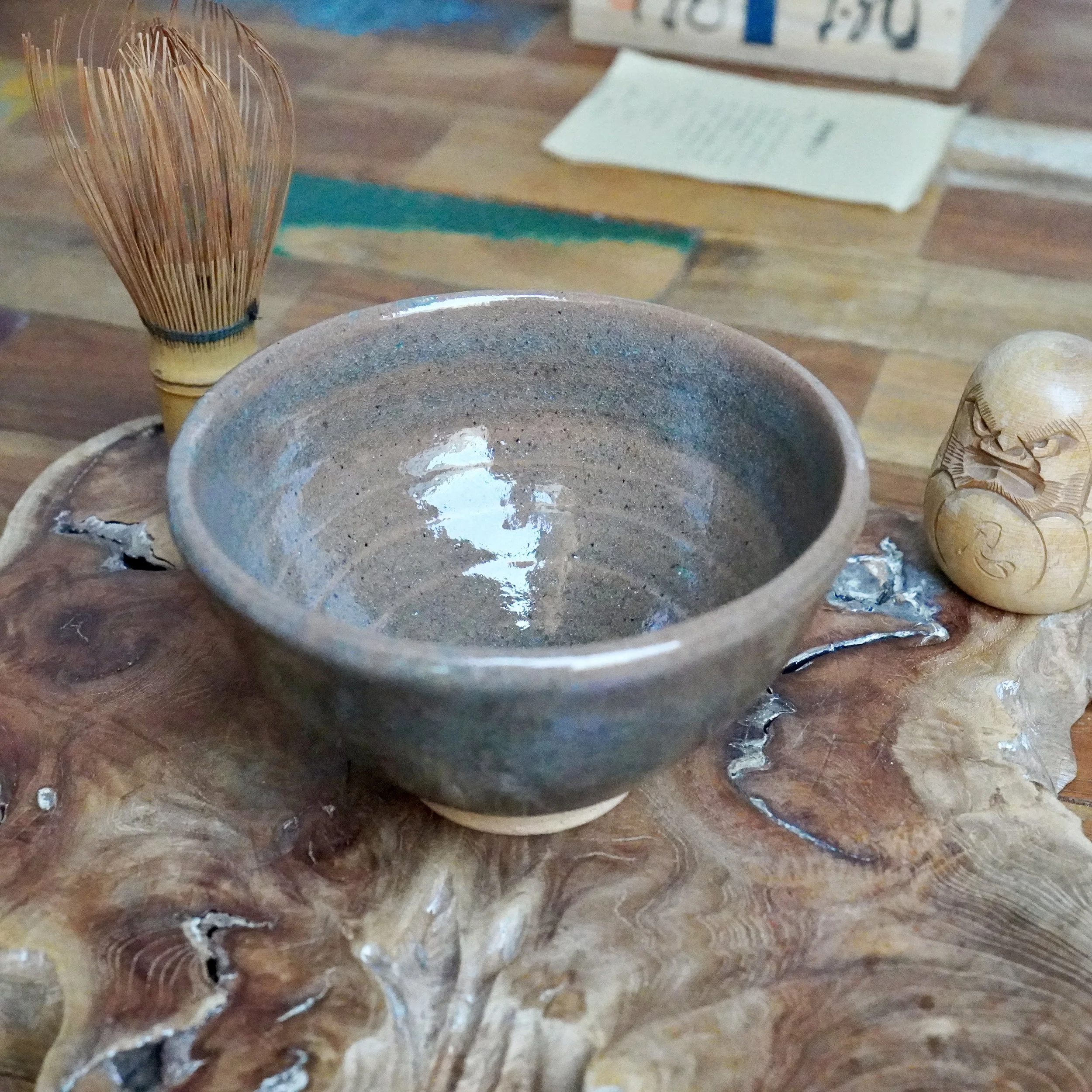 Mizushima Jirō's blue-grey speckled chawan, reflecting light, with a bamboo whisk and carved wooden figure on a textured wooden slab.