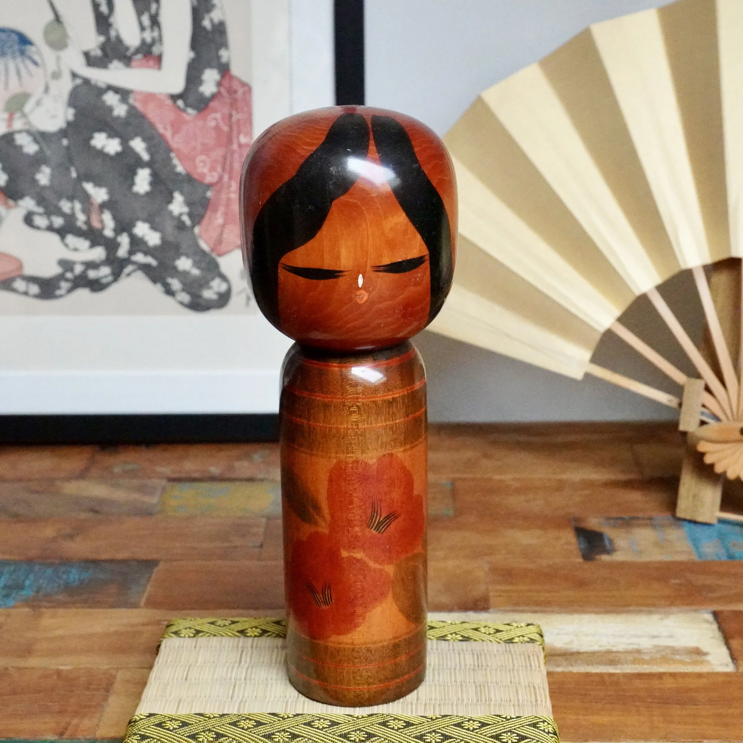 Tamura Noboru's Camellia Girl kokeshi doll, reddish-brown wood with black hair, heavy-lidded eyes, and red camellia flowers, on a woven mat.