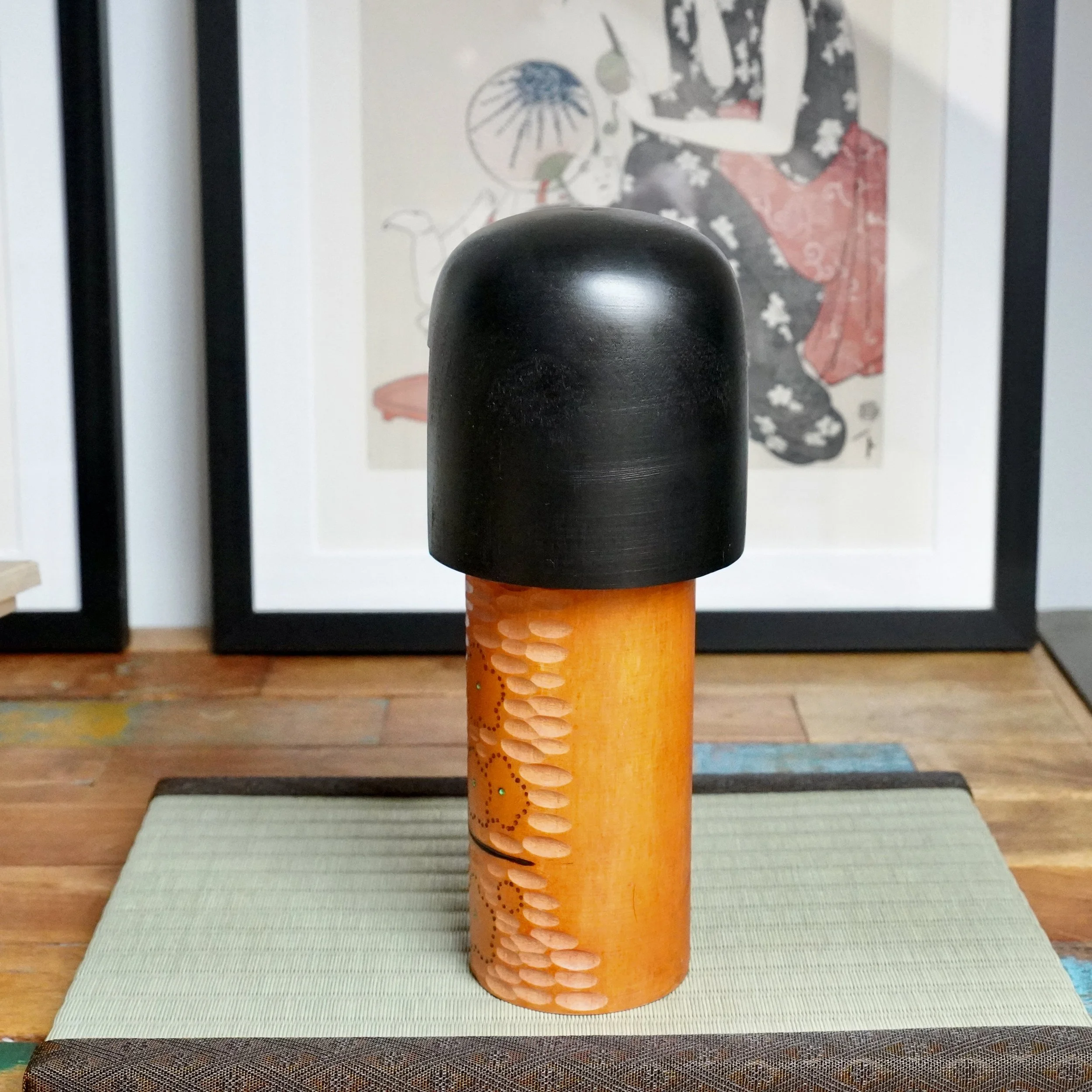 Kisaku sōsaku kokeshi, warm terracotta body with carved texture, painted ume blossoms, green dots, and black head, on a tatami mat.