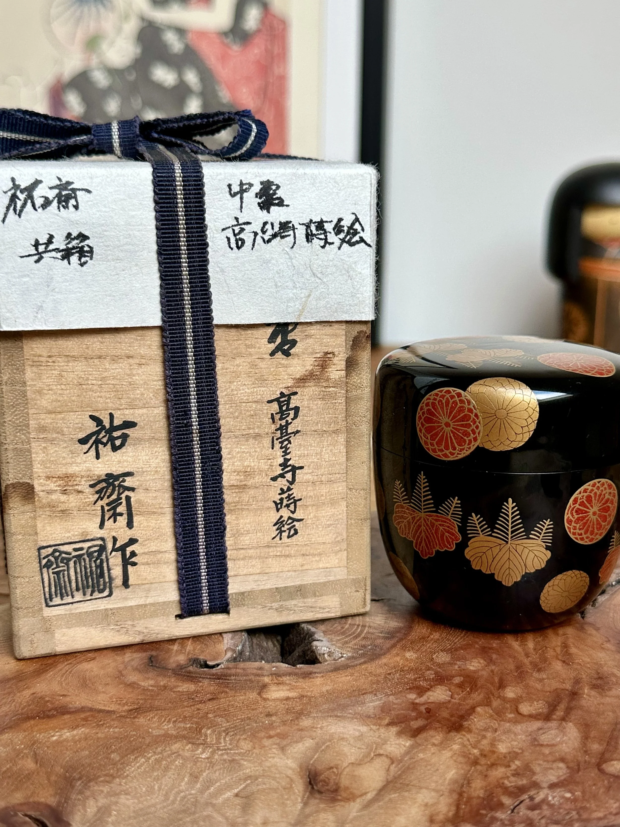 Black lacquered natsume tea caddy with gold and red makie crests, beside its wooden tomobako box with Hariya Yūsai's calligraphy and a blue ribbon.