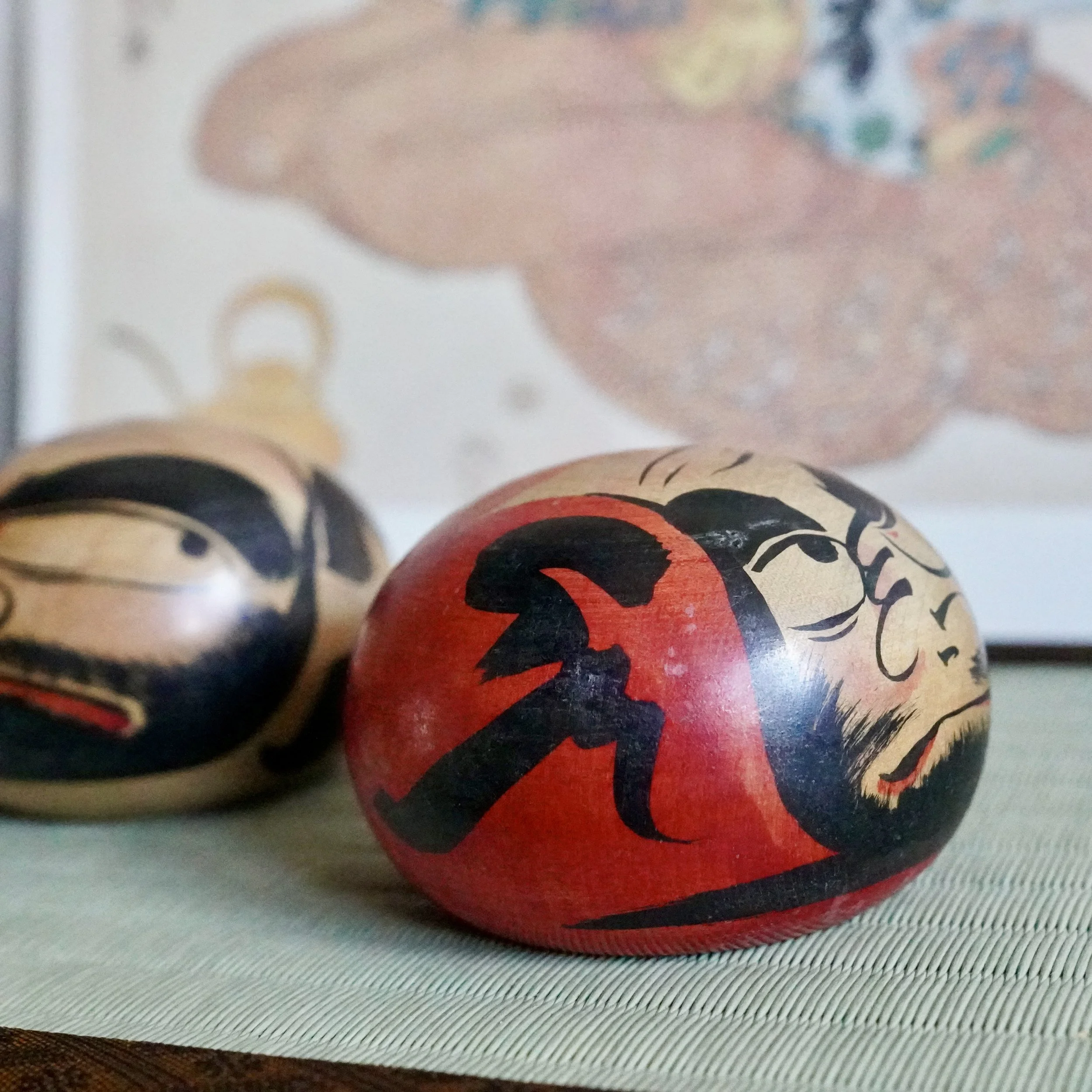 Two hand-painted daruma dolls with fierce expressions on a green woven mat. The front doll is red with black calligraphy; the other is natural wood.
