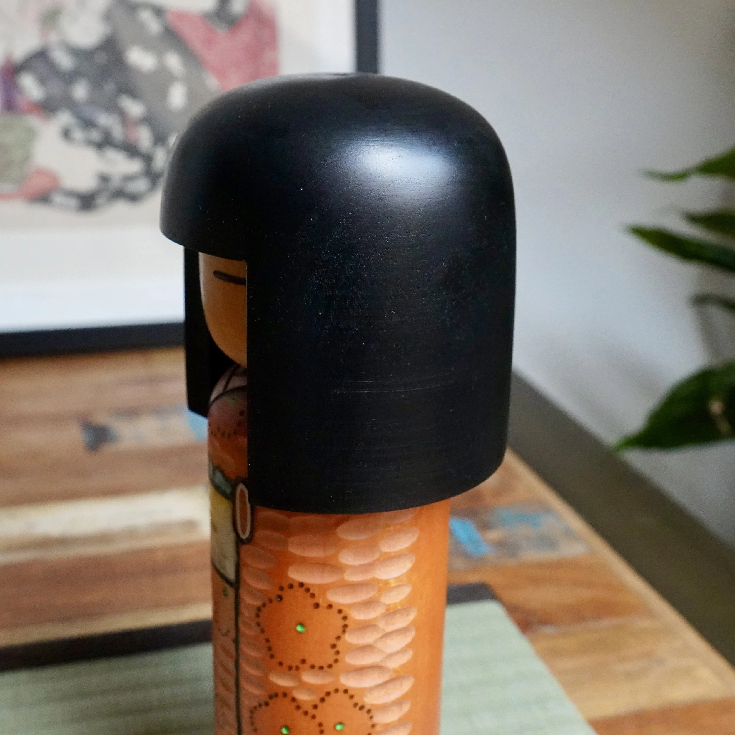 Kisaku kokeshi doll in side profile, with black hair covering a light brown head. Its orange kimono features carved indentations and green-centered ume blossom patterns.