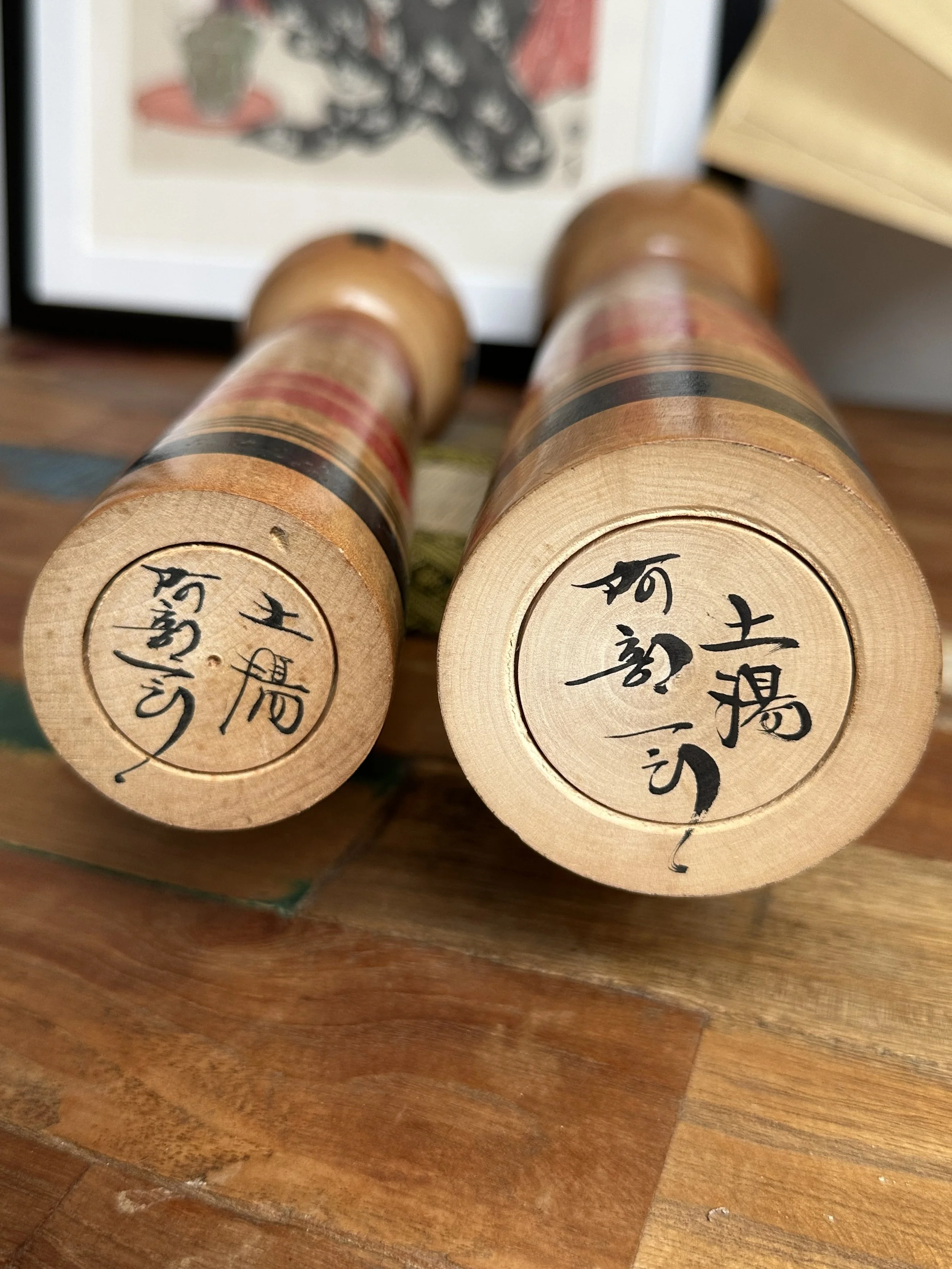 Two kokeshi doll bases by Abe Ichirō, showing carved black Japanese calligraphy signatures within circular grooves on natural wood.