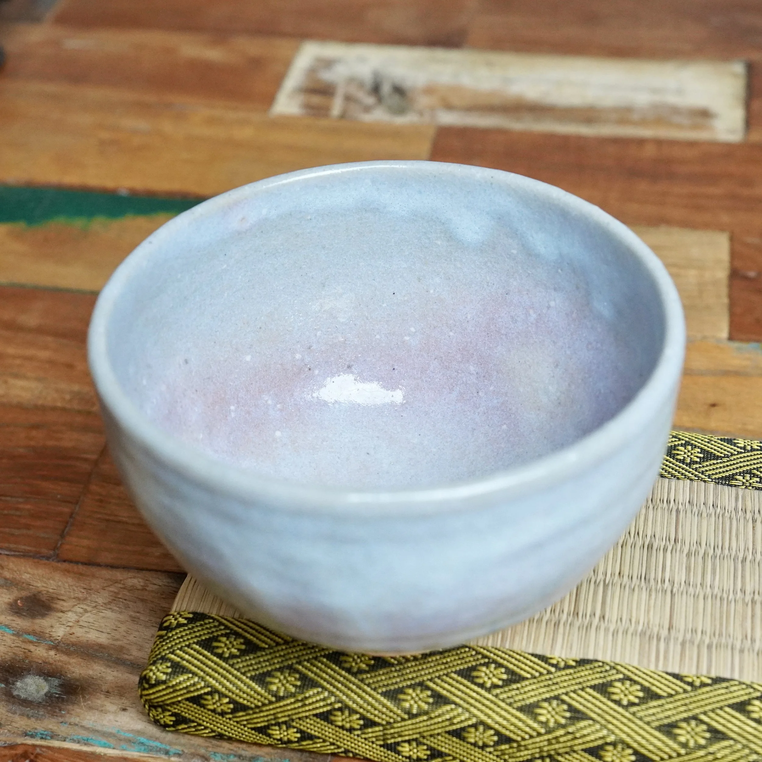 Moriwaki Fumitada's Hagi-yaki chawan, its blue-grey and lilac interior seen from above, rests on a patterned mat with green and yellow trim, over wood.