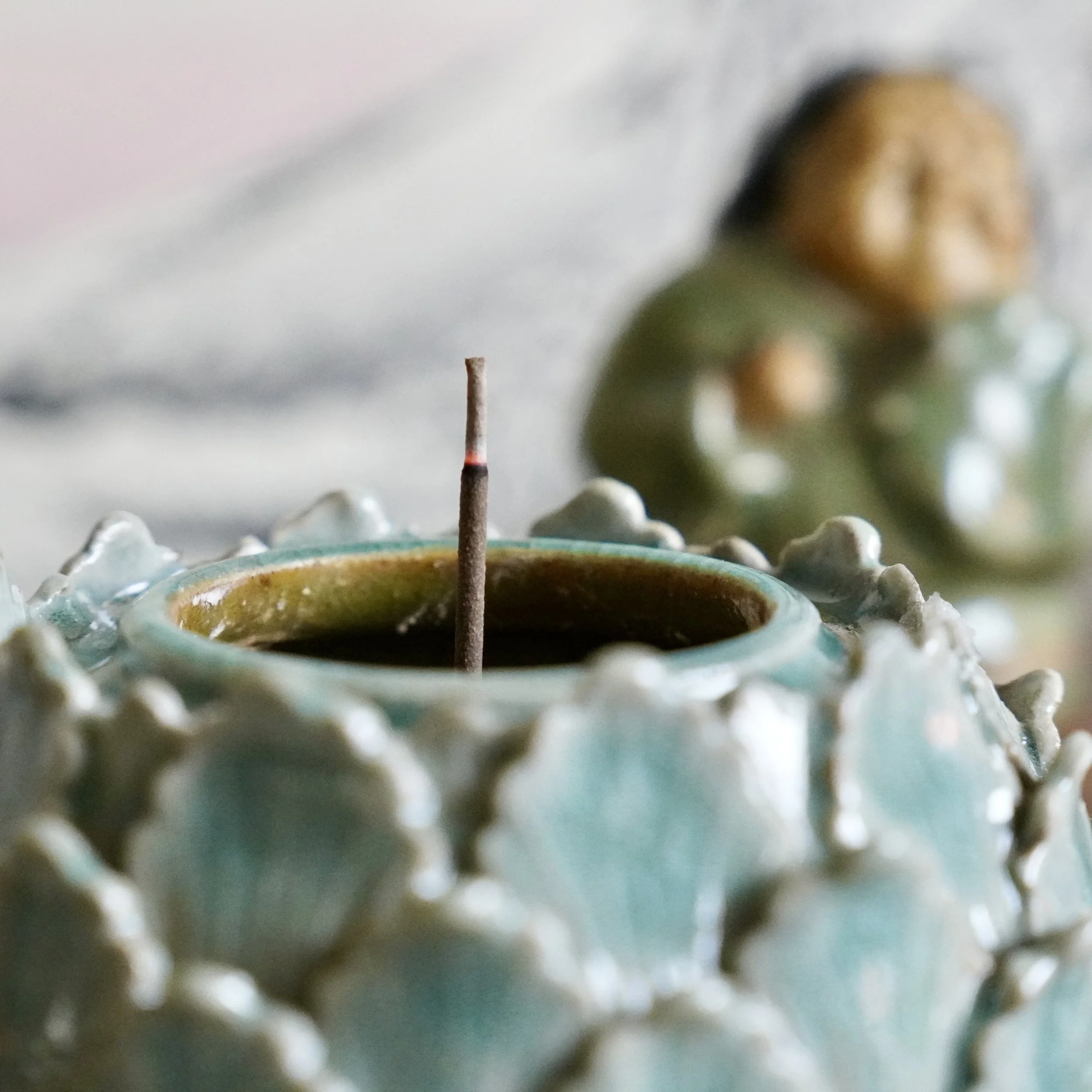Close-up view of incense lit with a tall, thin wick in the foreground and a blurred, golden-brown koro in the background.