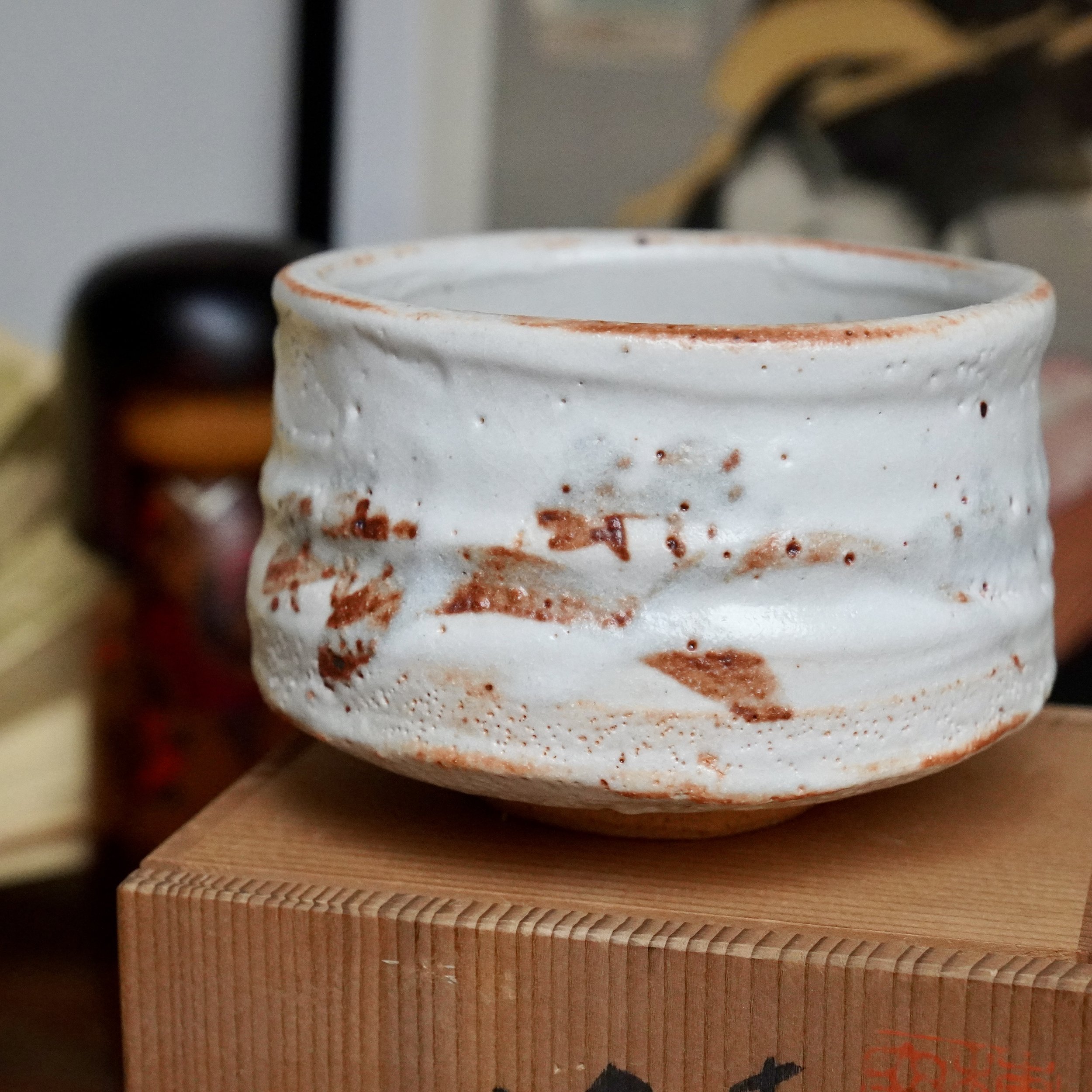 Handcrafted white Shino chawan with rust-orange splotches and an irregular rim, resting on a wooden tomobako with visible finger joints.