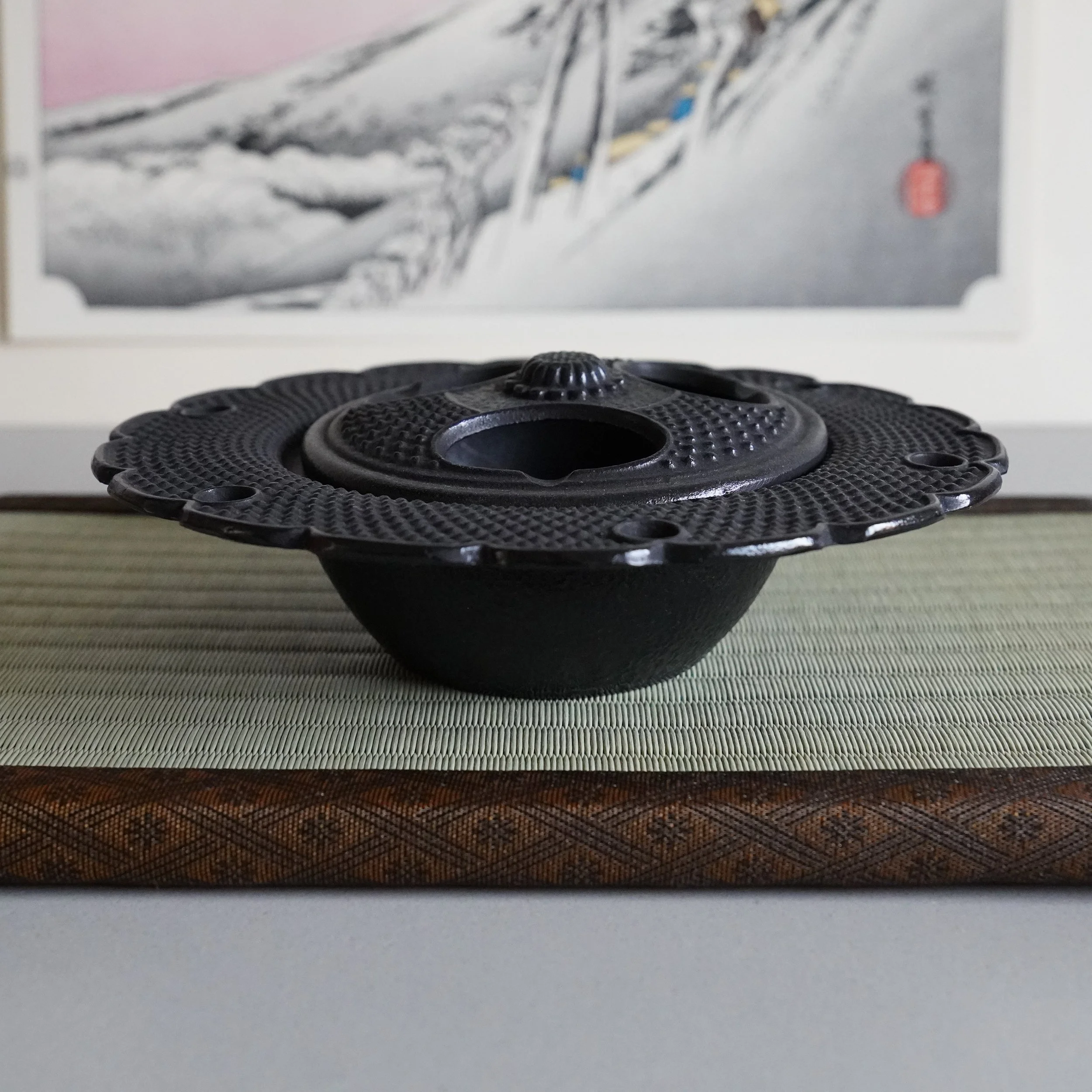 Side view of a Japanese cast iron kōro with sakura-shaped lid and arare hobnail texture, ukiyo-e print in background