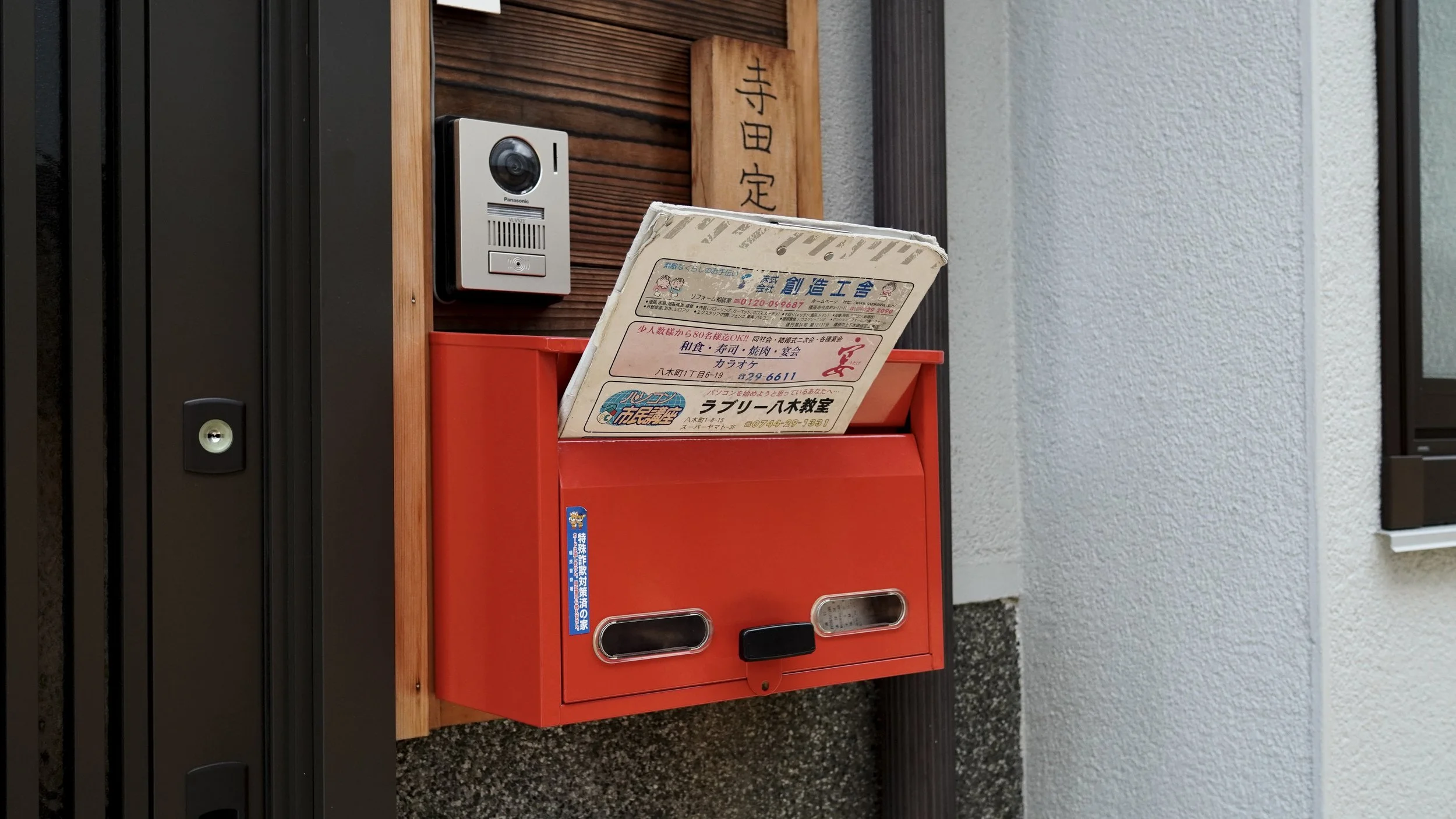 A red mailbox with a slot and a lock, mounted on a wooden panel, with Japanese newspaper or pamphlet sticking out of it. A doorbell and a wooden plaque with Japanese characters are nearby.