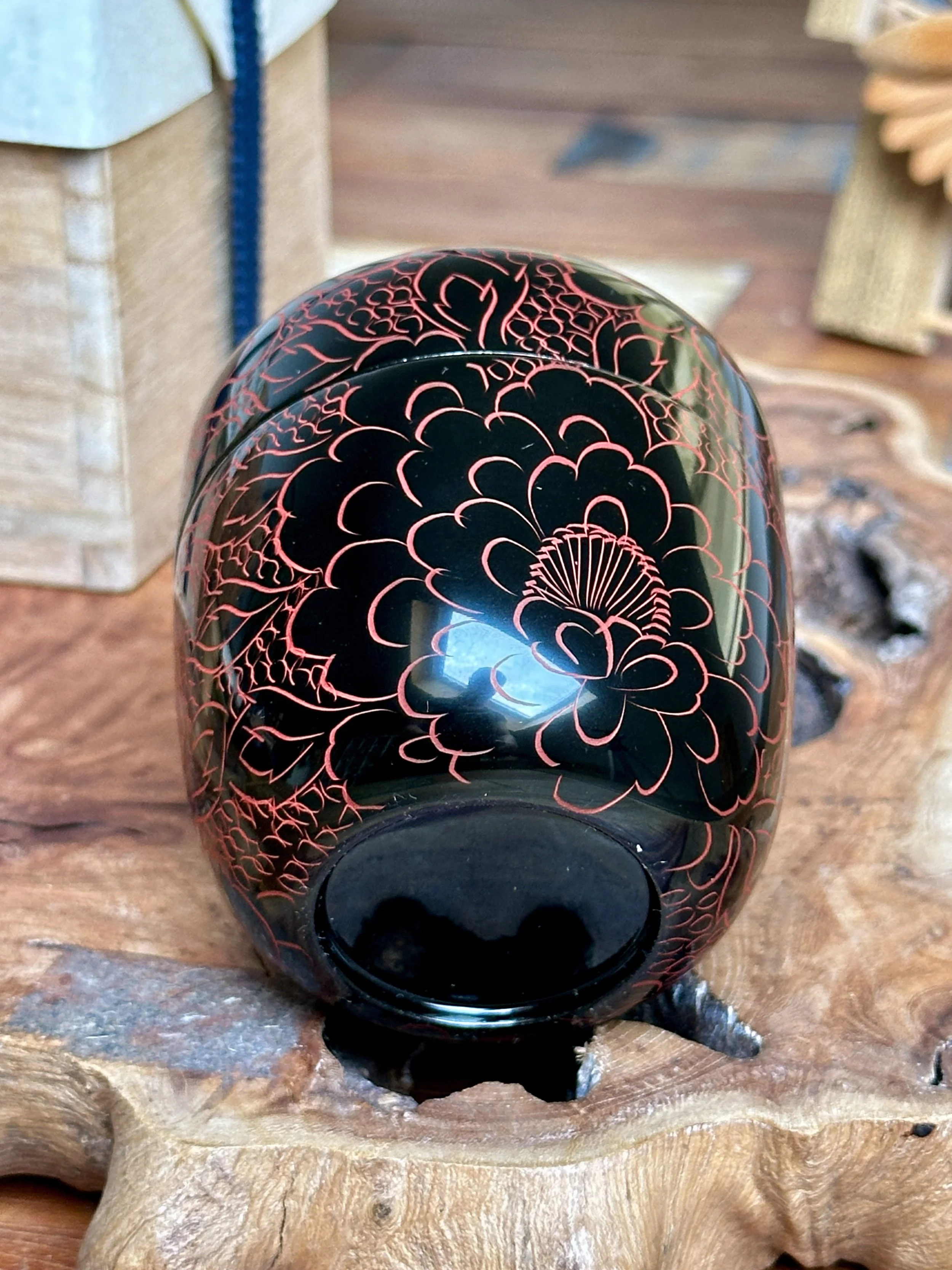 Rounded, glossy black tea caddy by Fujiwara Seiu, featuring engraved vermilion peony blossoms and leaves, resting on a wooden surface.