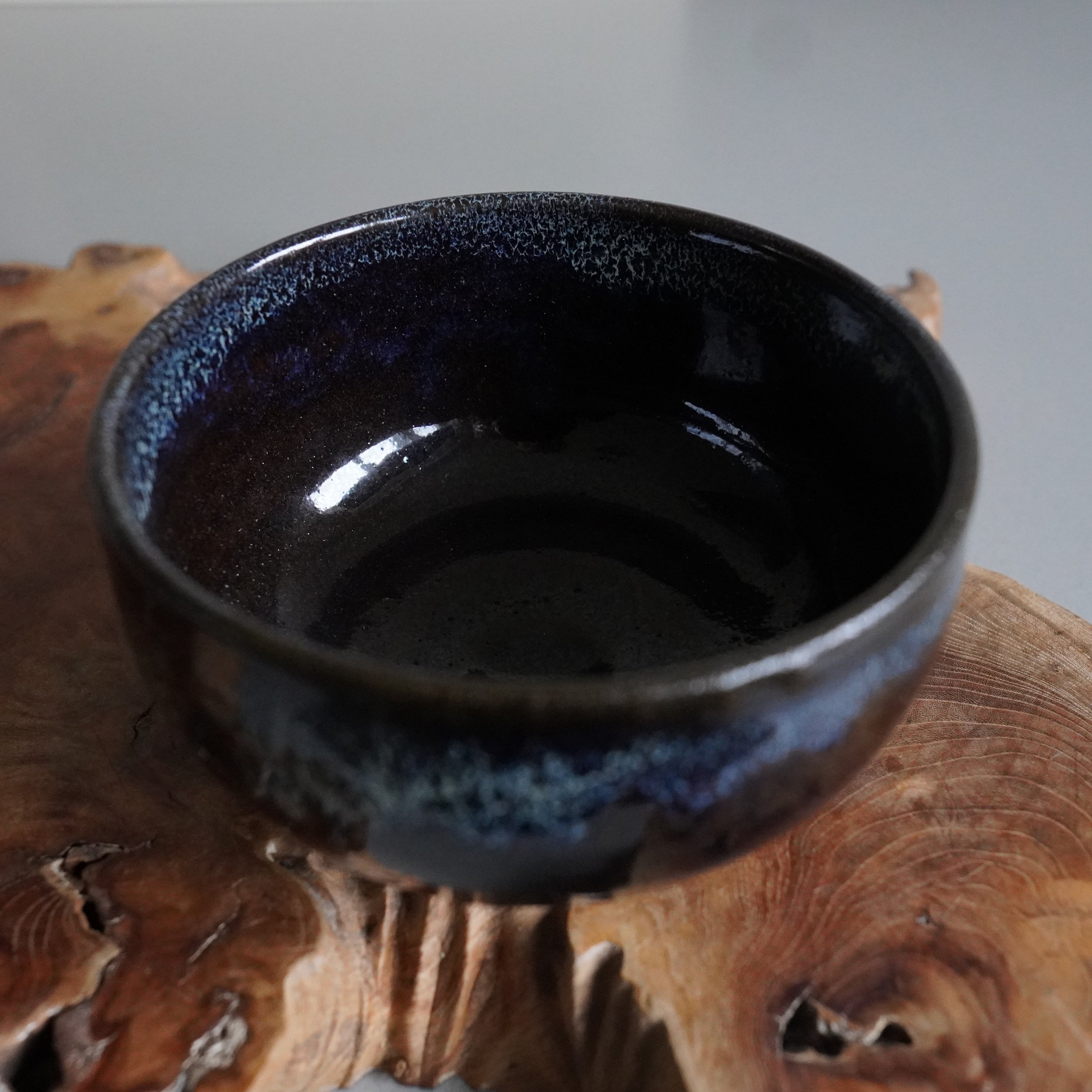 Dark ceramic matcha bowl by Rokuhō, with mottled blue-black glaze and reflective interior, resting on a textured wooden slab.