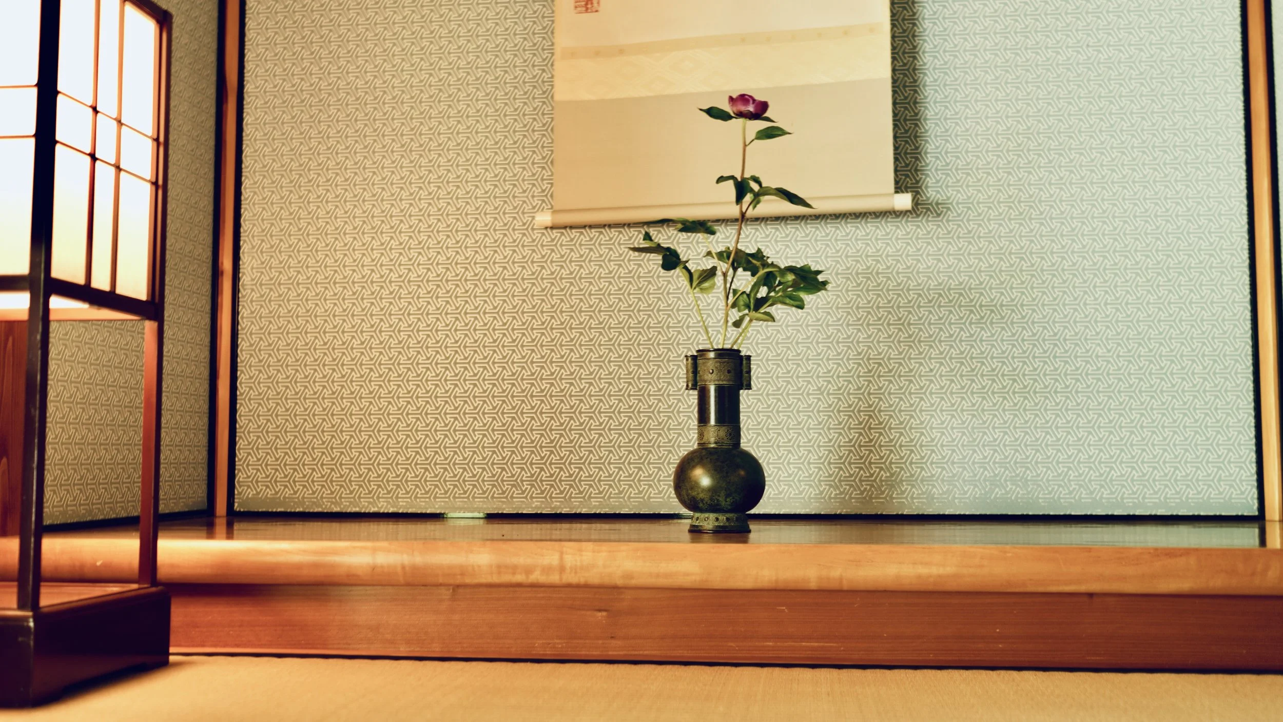 Japanese tea room tokonoma alcove with a dark bronze ikebana vase holding a single rose stem, and a kakejiku hanging scroll with ink painting above. Tea ceremony room at Kenroku-en garden, Kanazawa.