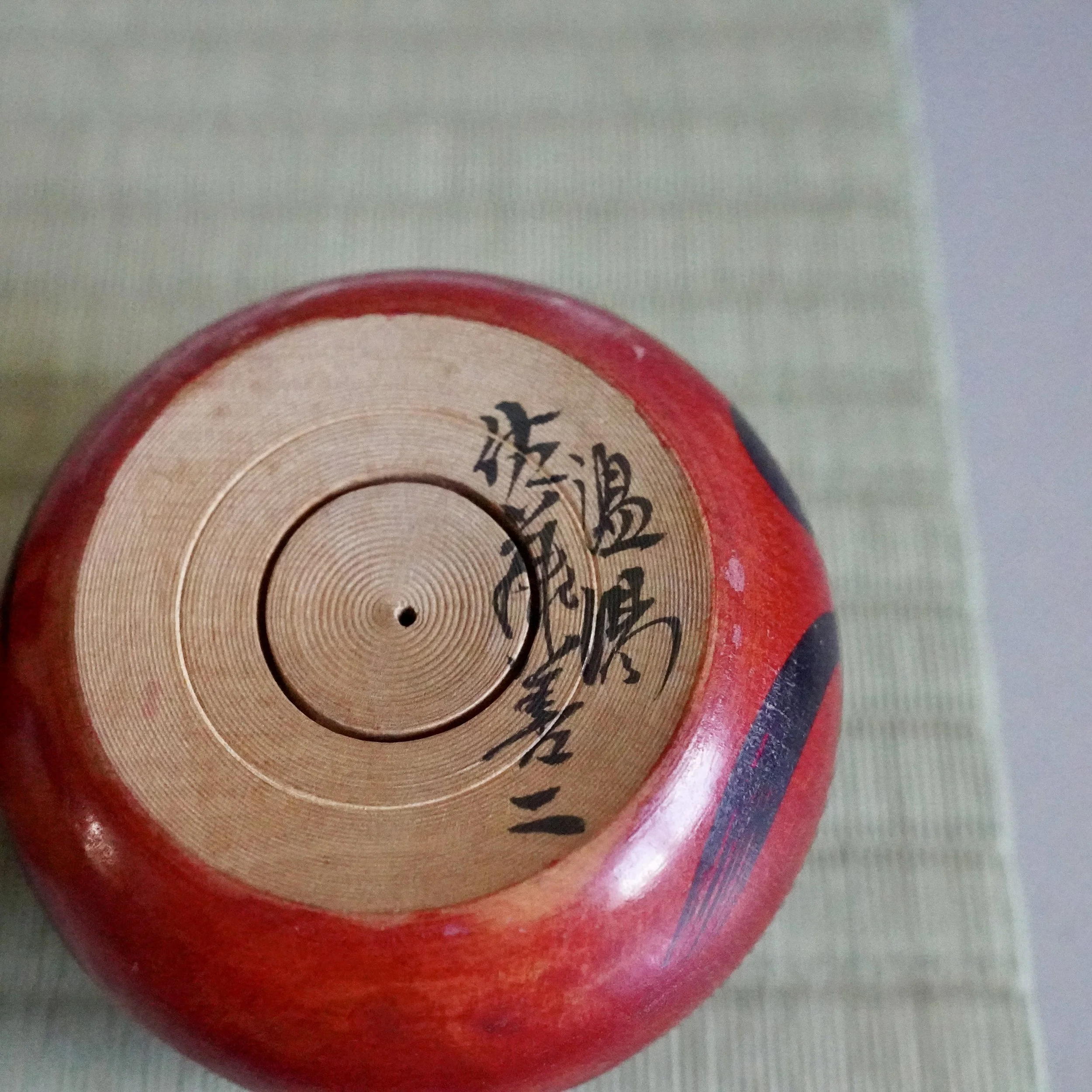 Underside of a red Tsugaru daruma, showing its circular wooden base with concentric rings. Black calligraphy, the signature of Satō Zenji and Nuruyu, is painted on the natural wood.