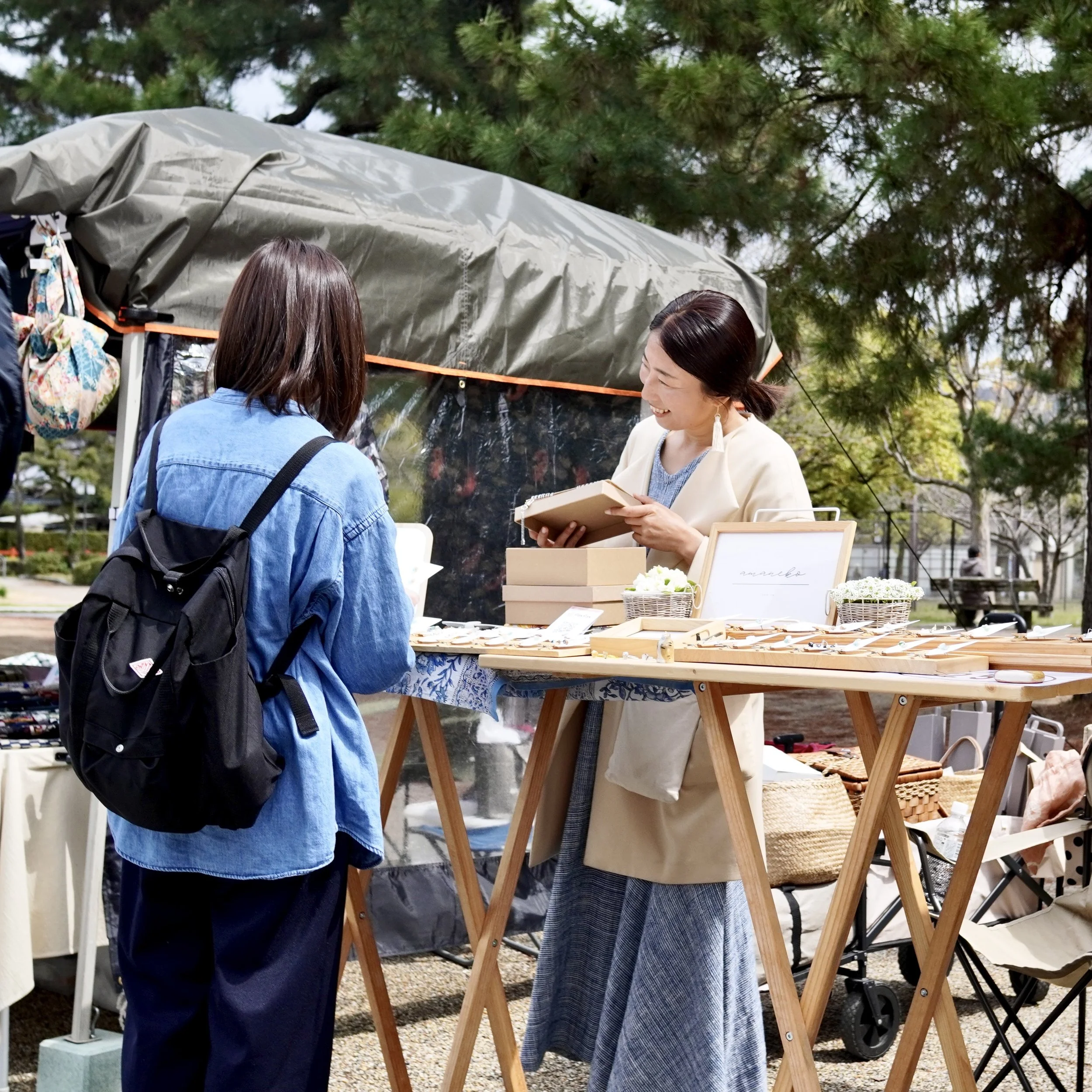 A cheerful Japanese seller at an outdoor market interacts with a customer at a table filled with small handmade boxes and accessories during a sourcing trip.