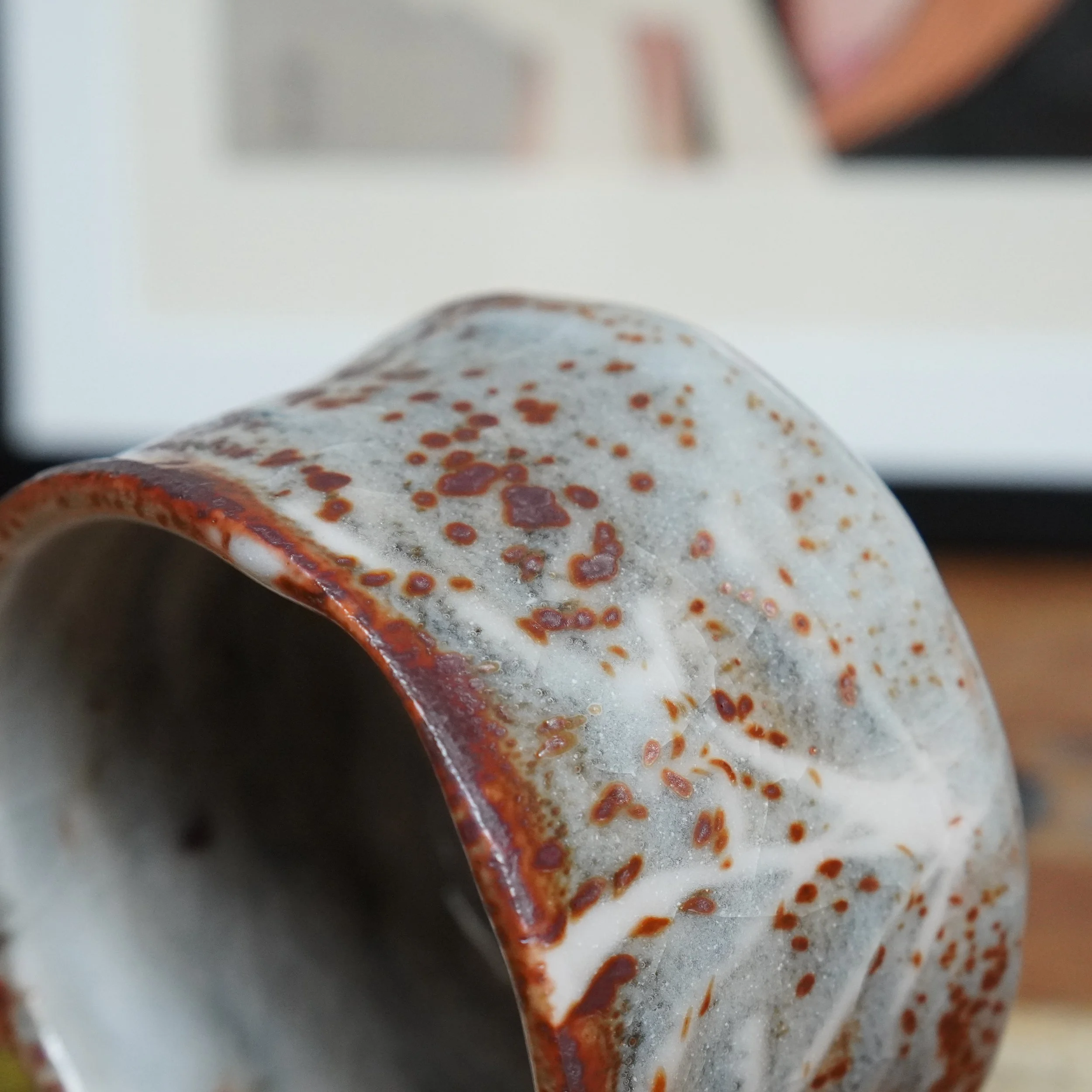 Angled close-up of a Shino chawan. Its pale grey glaze is dotted with reddish-brown speckles and crossed by a white branch-like pattern, with a dark red rim.