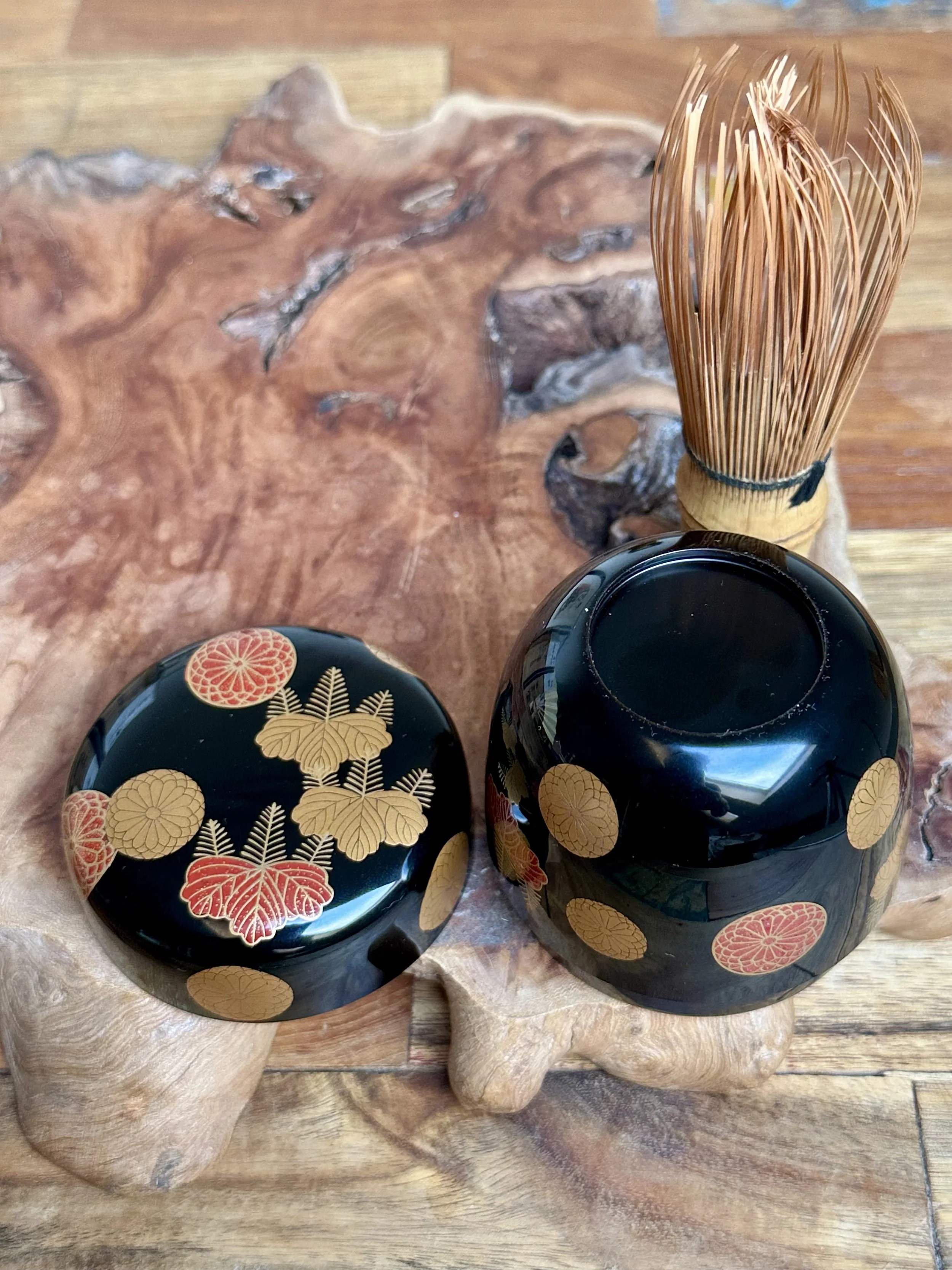 Yūsai's black lacquered natsume tea caddy with gold and red chrysanthemum & paulownia crests, beside a matcha whisk on wood.