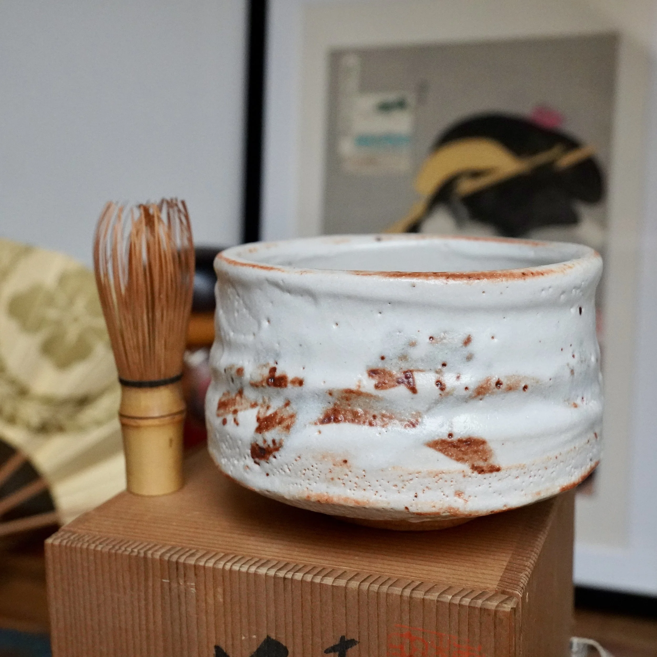 Shino chawan with white glaze and rust-orange blush, a bamboo whisk, and its carved wooden tomobako. Visible from front.
