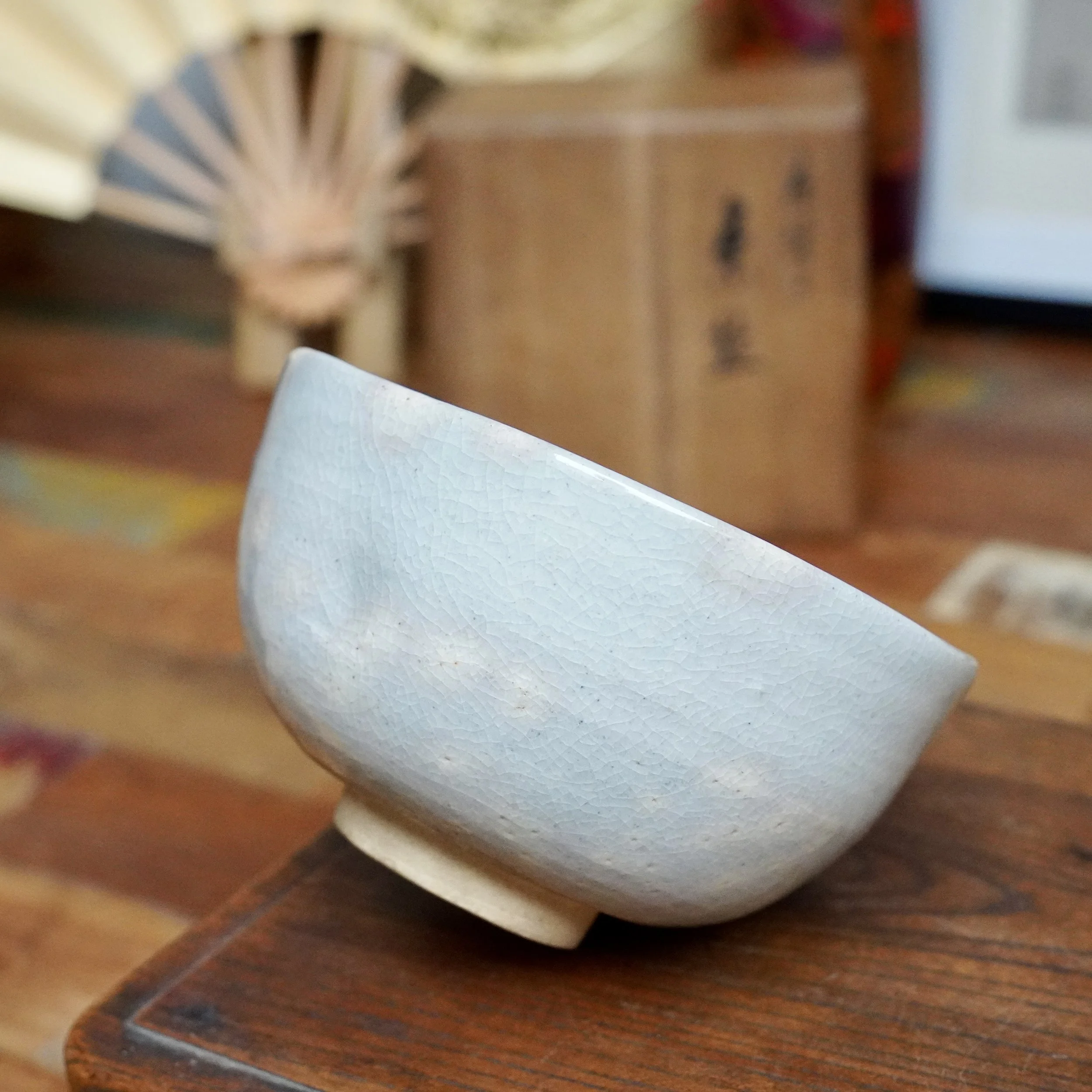 Nagasawa Minoru's Izushi chawan, soft grey-blue crackle glaze with scattered pink blush, angled on a dark wooden table.