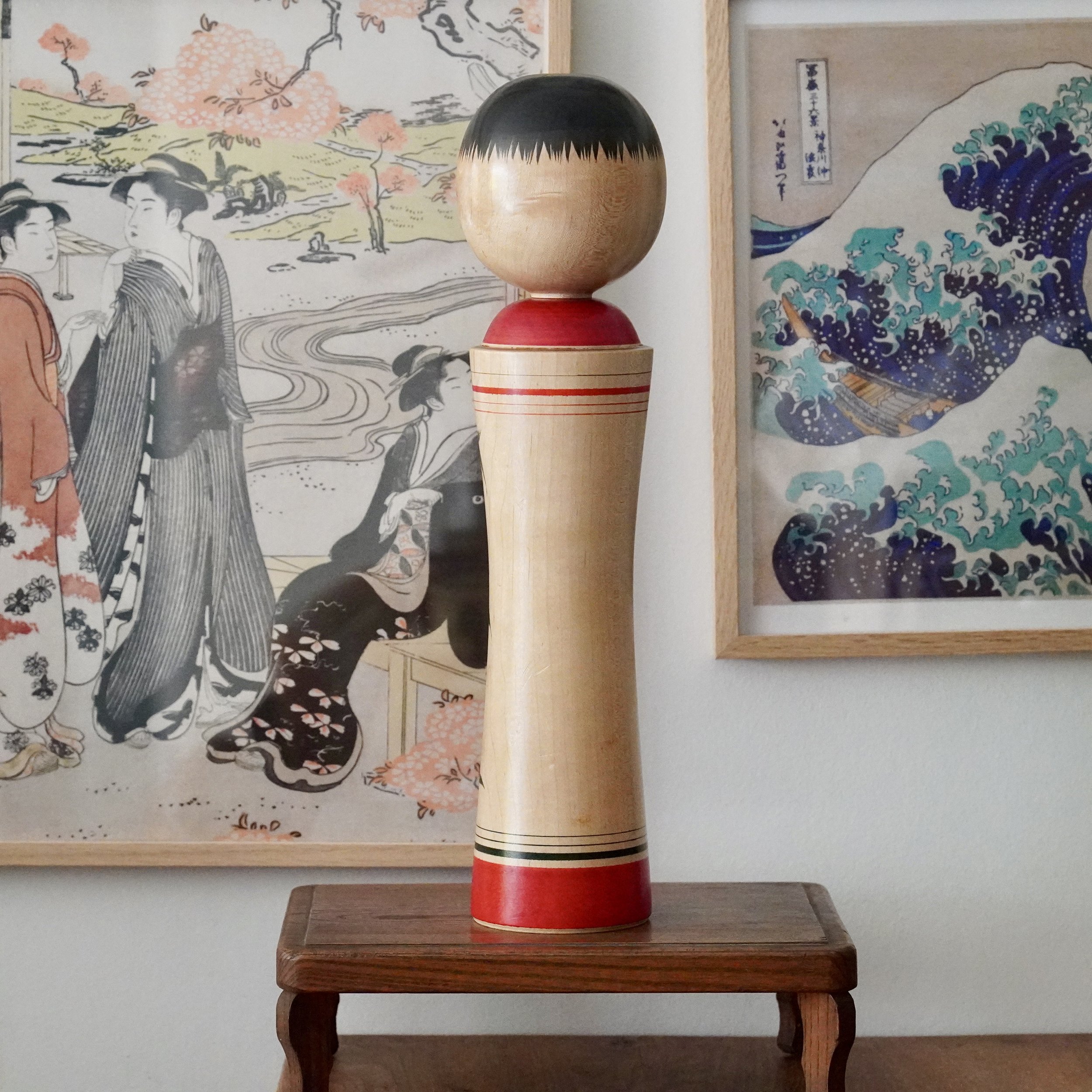 Izu Mamoru kokeshi doll, light wood, black fringed hair, red collar and base, with red and black stripes. On a dark wood table, flanked by two Japanese prints.