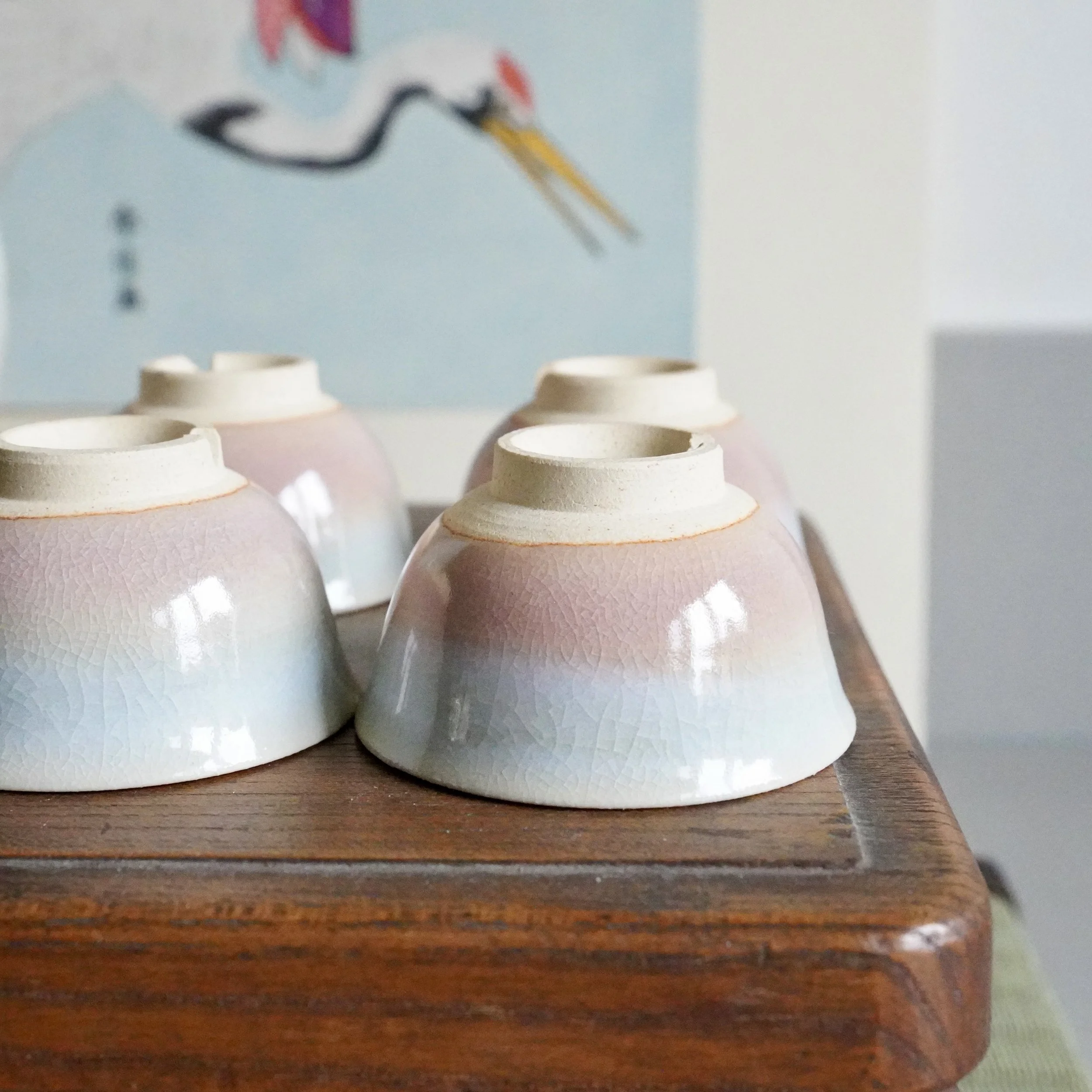 Four Hagi-yaki ochoko cups, upside down on a wooden surface, displaying a gradient glaze from blush pink to pale blue-grey with fine crazing.