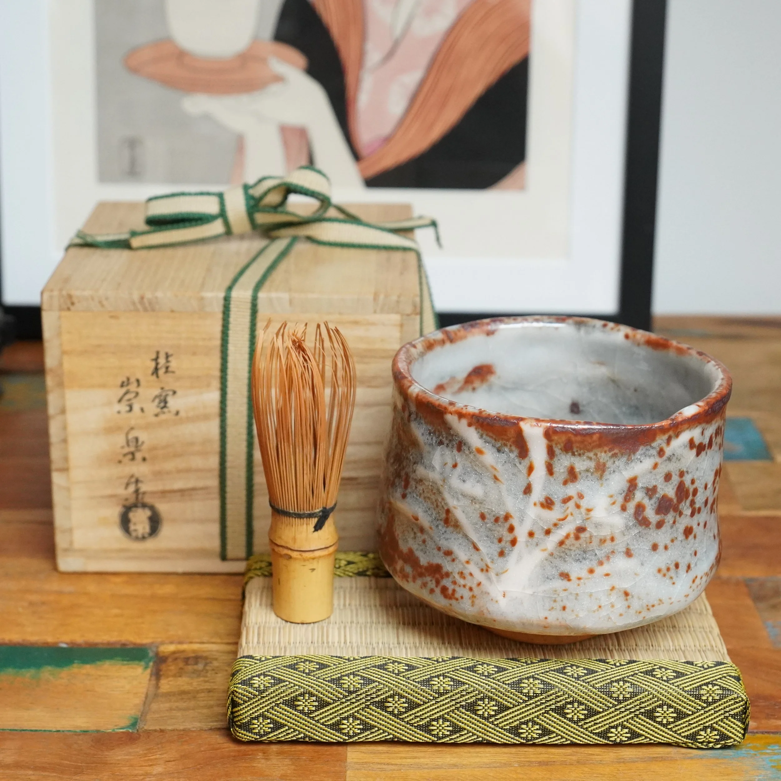 Shino chawan by Katsura kiln with white and red speckled glaze and crazing, a bamboo whisk, and tomobako with ribbon on a mat.