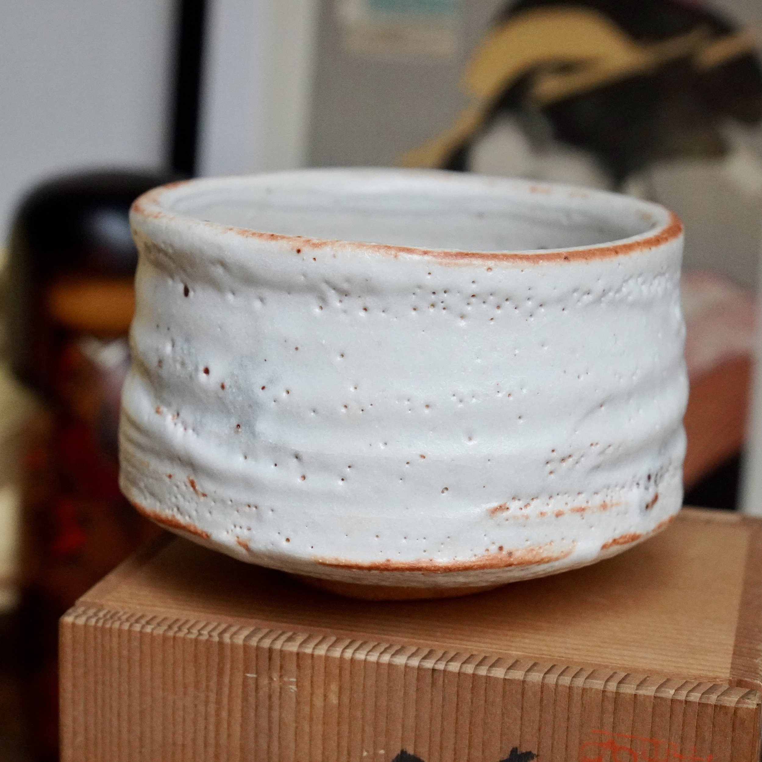 Textured white Shino chawan with rust-orange blush at the rim and base, resting on a light brown wooden box with visible finger joints.