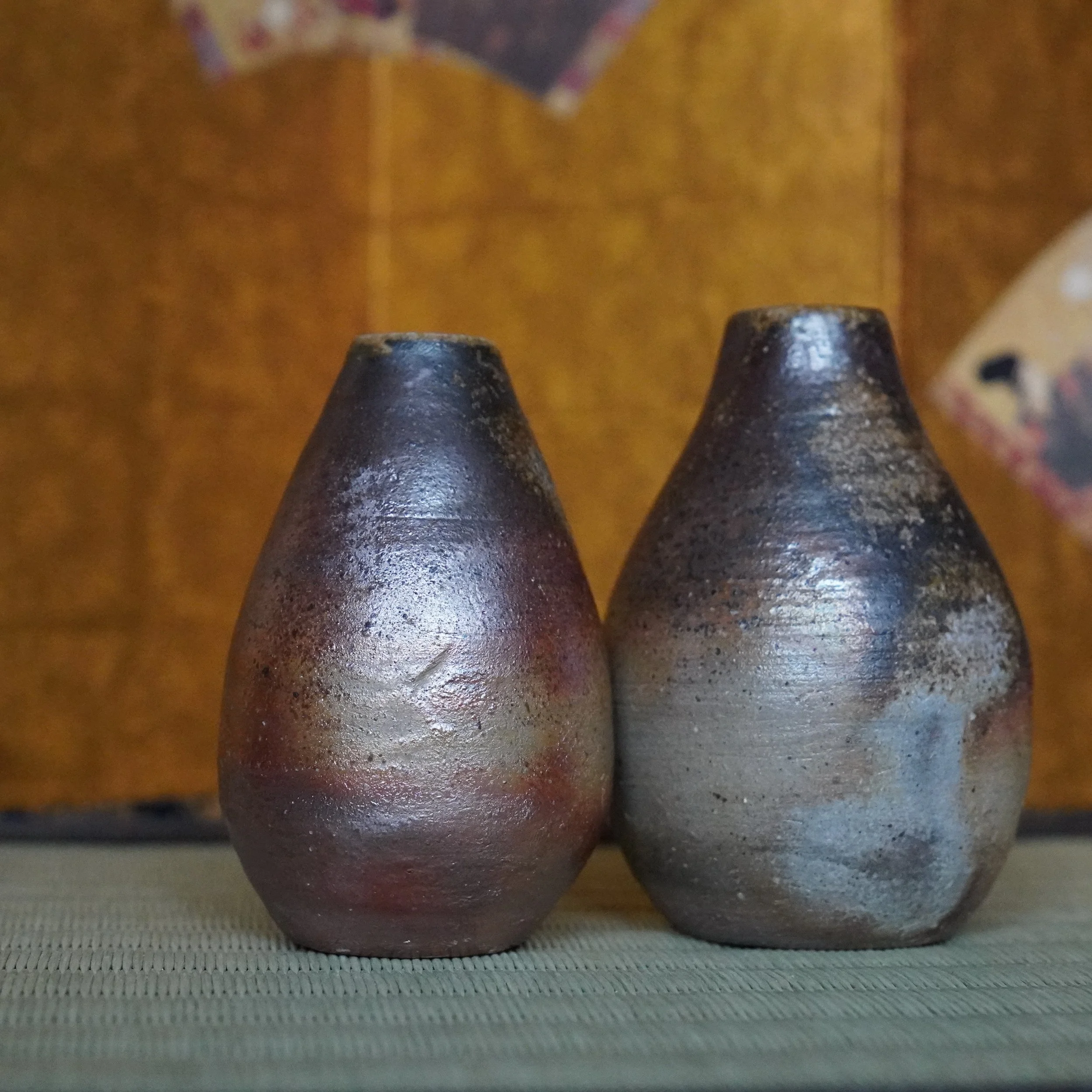 Shibaoka Tōsendō Bizen-yaki vases, a dark reddish-brown piece with silver highlights and a lighter grey-brown piece, both rustic and textured, stand on a woven mat before a blurry golden background.