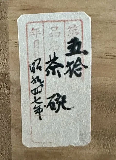 Off-white paper label with black handwritten Japanese characters, including '茶盌' (tea bowl), '昭和四十七年' (Shōwa 47), and '鹿峰' (Rokuhō), framed by a red border on a light brown surface.