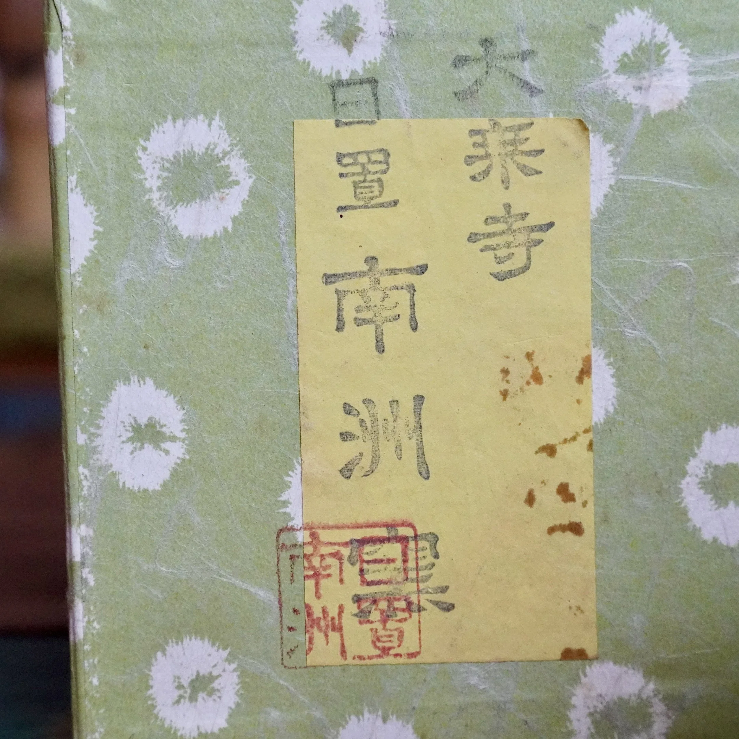 Yellow box label with vertical black Japanese characters "Daijō-ji" and "Hioki Nanshū", and a red seal. It's on green, fibrous paper with white, dandelion-like patterns and brown spots.