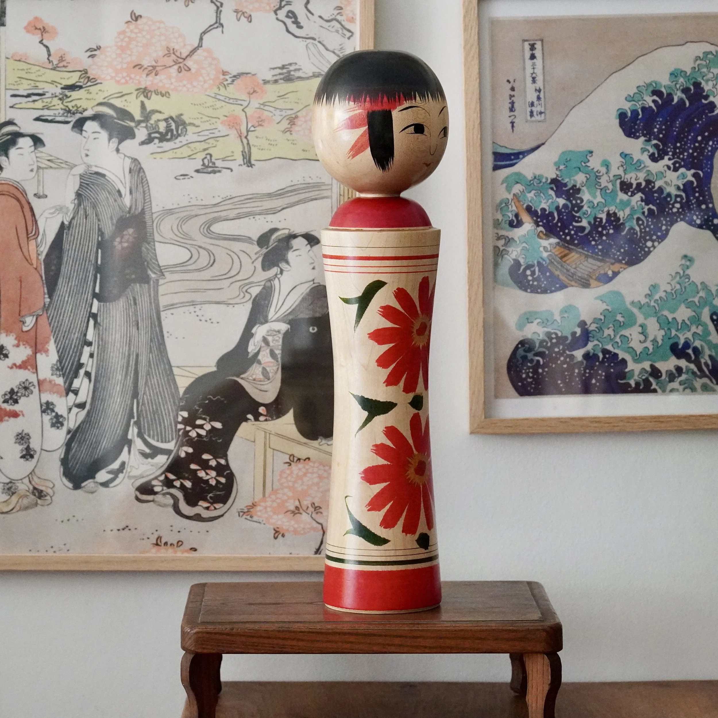 Izu Mamoru Ginzan kokeshi, pale wood body, red chrysanthemums, red trim, black bob, simple face. On wooden stand, flanked by Japanese prints.