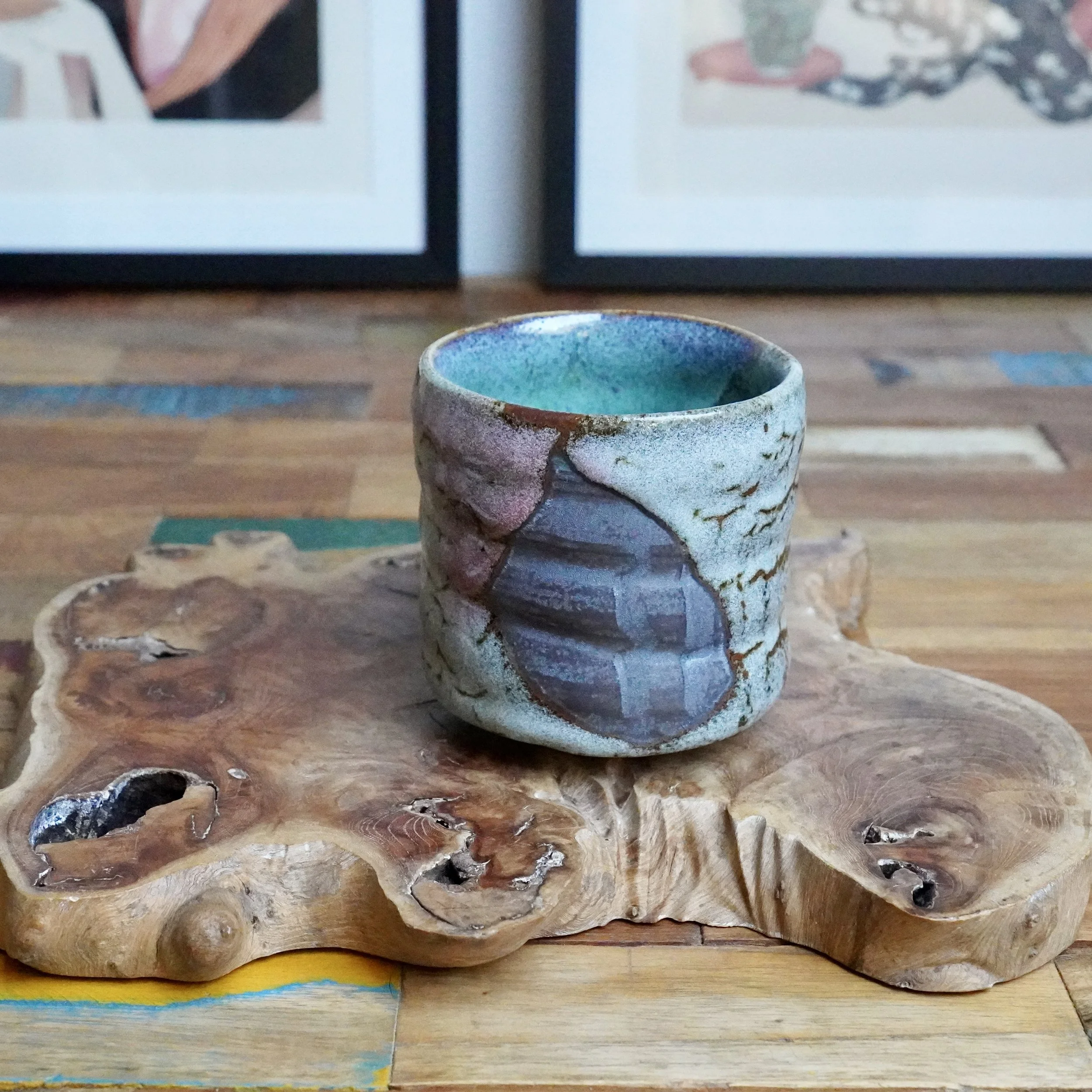 Hand-thrown tsutsu chawan with clear turquoise interior, layered purple, grey, and brown exterior glazes with a ridged design, on a rustic wooden slab.