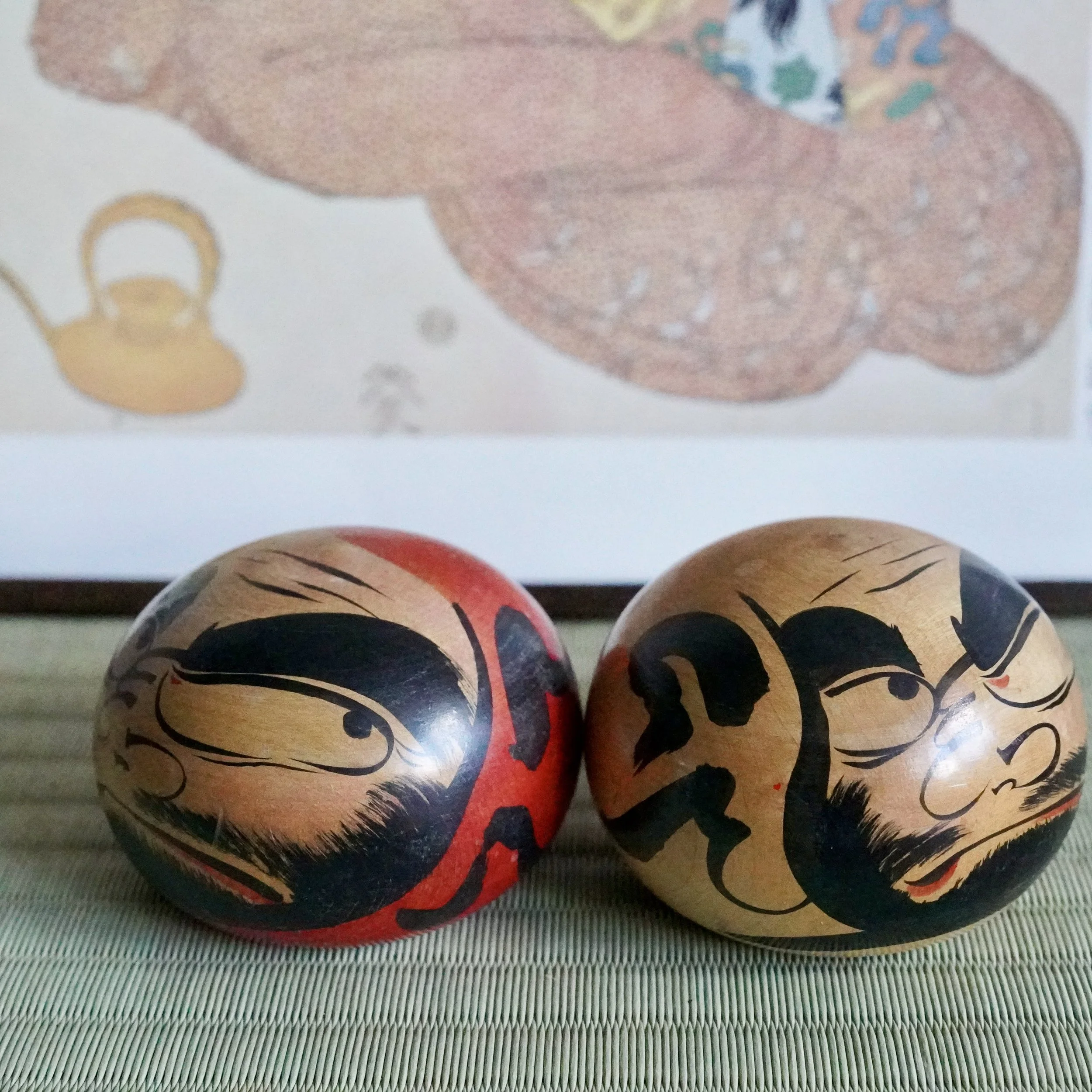 Two round Tsugaru daruma by Satō Zenji, one red and one wood-toned, with black fierce faces and red accents, sit on a woven mat.