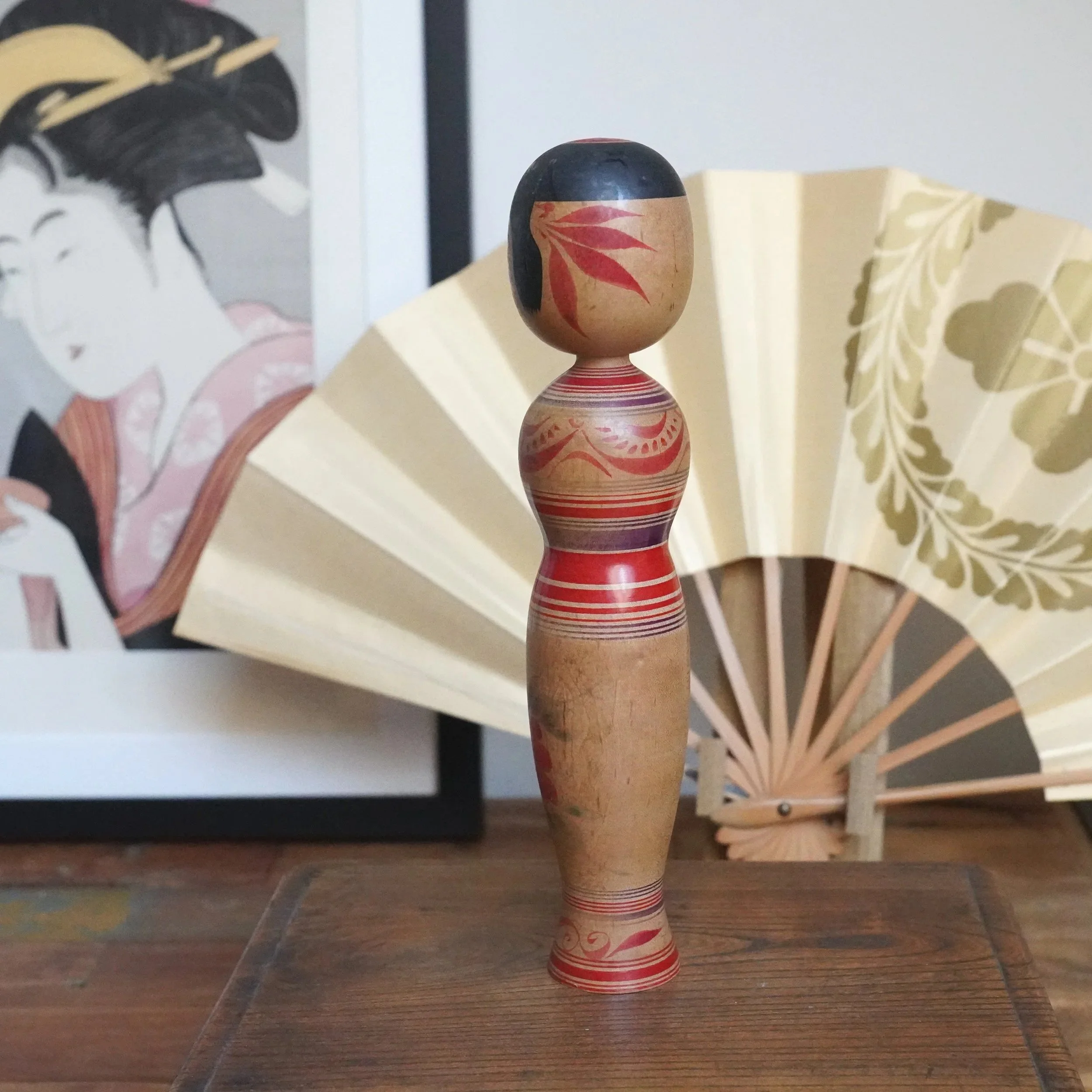 Sasamori Junichi Tsugaru kokeshi, hourglass-shaped with a black head, red peony and striped decoration. A Japanese print and cream fan are behind.