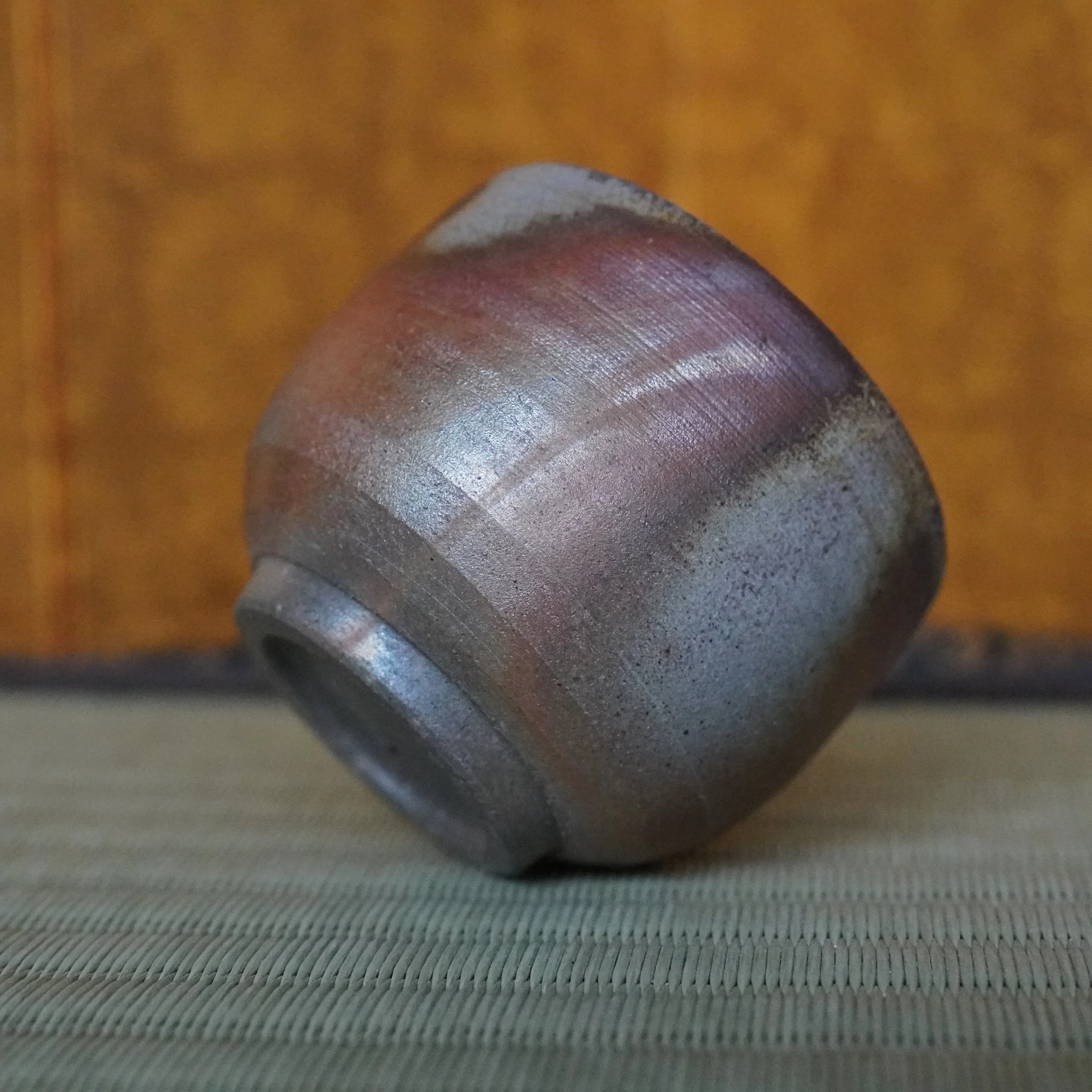 Tilted Bizen-yaki guinomi by Yamamoto Sōshū, featuring prominent reddish-purple hidasuki marks on its textured, unglazed body. It rests on a woven mat against an orange background.